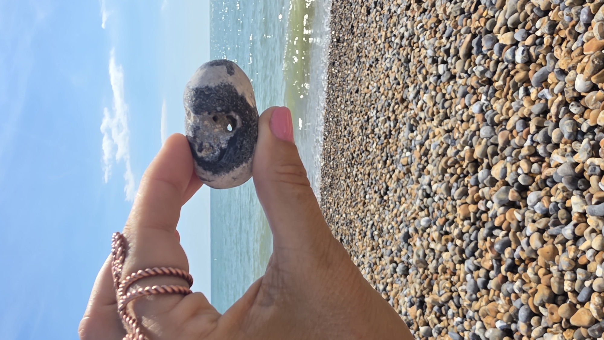A hand, adorned with a coiled copper ring and bright pink nail polish, holds a pebble with a hole through it in the foreground. The pebble is grey, white, and black. The background features a pebble beach, the blue sea, and a clear blue sky with scattered white clouds. The scene is consistent with a sunny day at the beach in St Leonards, United Kingdom.