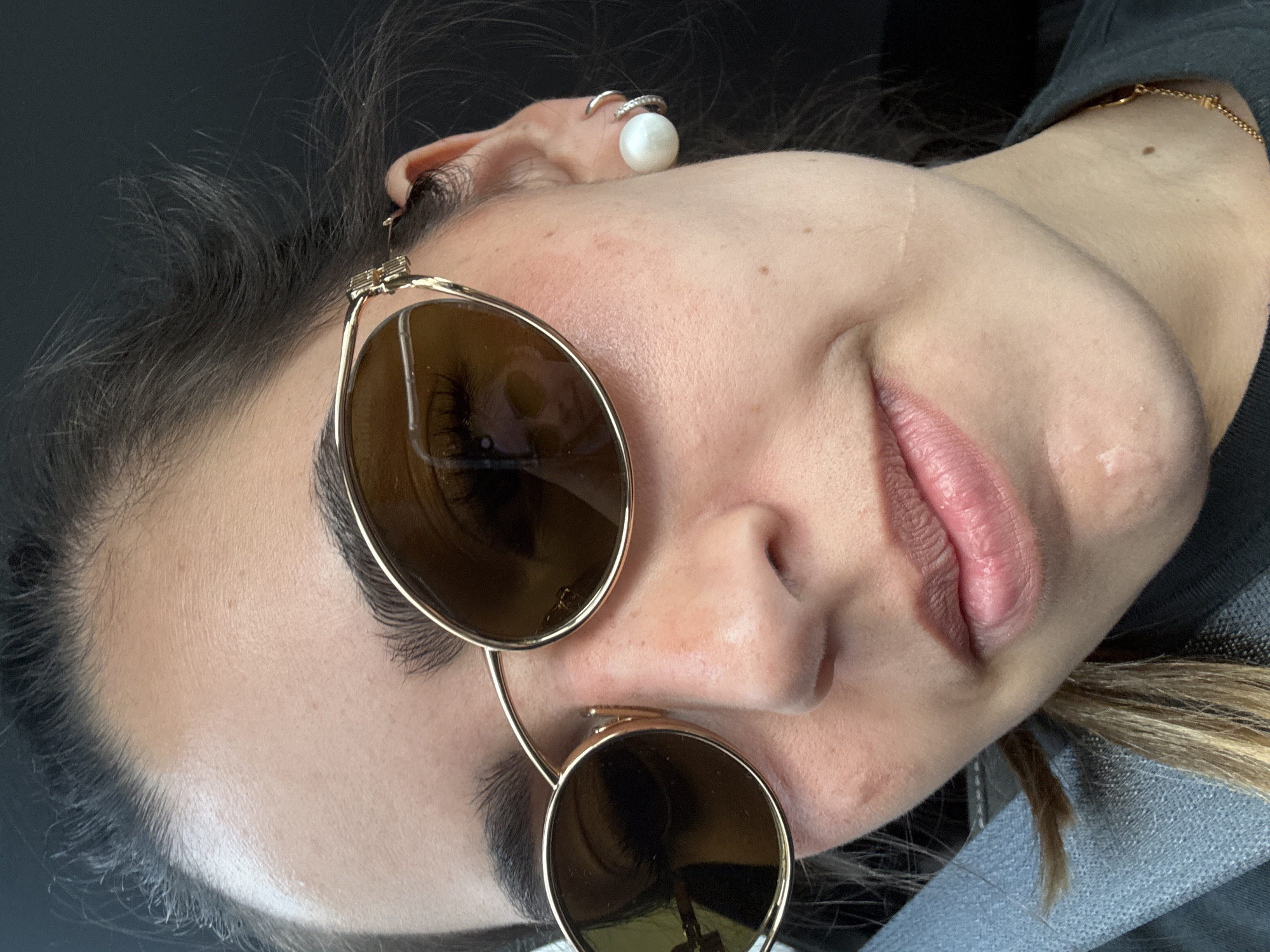 A close-up image of a light-skinned individual wearing gold-rimmed round sunglasses. The person has dark hair and is wearing two earrings in the left ear: a cuff earring near the top and a pearl stud in the lobe. A gold chain is visible on the neck. The person's lips are slightly parted and appear pink. The background is dark and out of focus. It is not possible to determine the specific location in Live Oak, United States of America, or any actions or interactions without further context.
