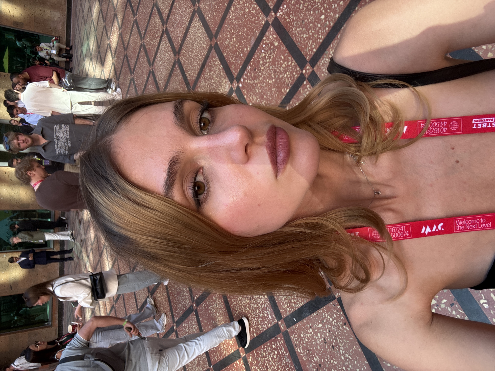 The image is a selfie of a woman with light brown hair, wearing a lanyard with "STBET PARTNERS" printed on it. The background shows an outdoor public area with a tiled floor of reddish-brown and black. Other people are walking around in the background, appearing slightly blurred due to the selfie's focus on the woman. The lanyard also says "Welcome to the Next Level" and has the numbers "40.16724" and "44.500/4" printed on it. Based on the lanyard of a partner organization of Yerevan, the location appears to be in the city of Yerevan, Armenia.