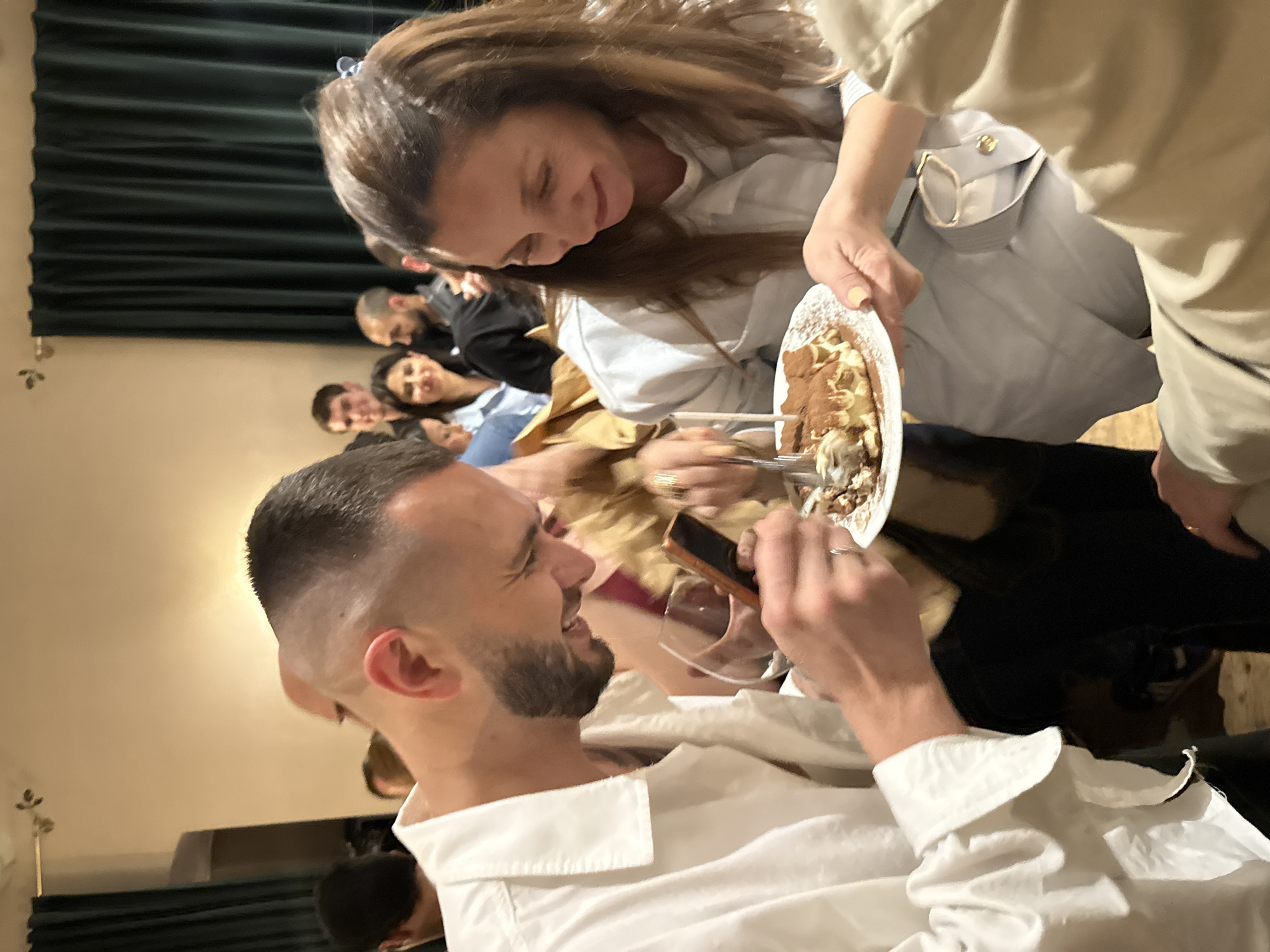 A horizontally-oriented medium shot captures a group of people indoors, possibly at a gathering. In the foreground, a man with a short, faded haircut and a trimmed beard, dressed in a white shirt, is smiling as a woman holds a plate of dessert towards him. She has long brown hair and is wearing a light-colored jacket. She is offering him a spoonful of the dessert. The man is holding a wine glass with his left hand and a phone with his right hand. Several other people are visible in the blurred background, seated and appearing to be part of the same group. A green curtain is visible behind them. The location is likely indoors and appears to be Yerevan, Armenia.