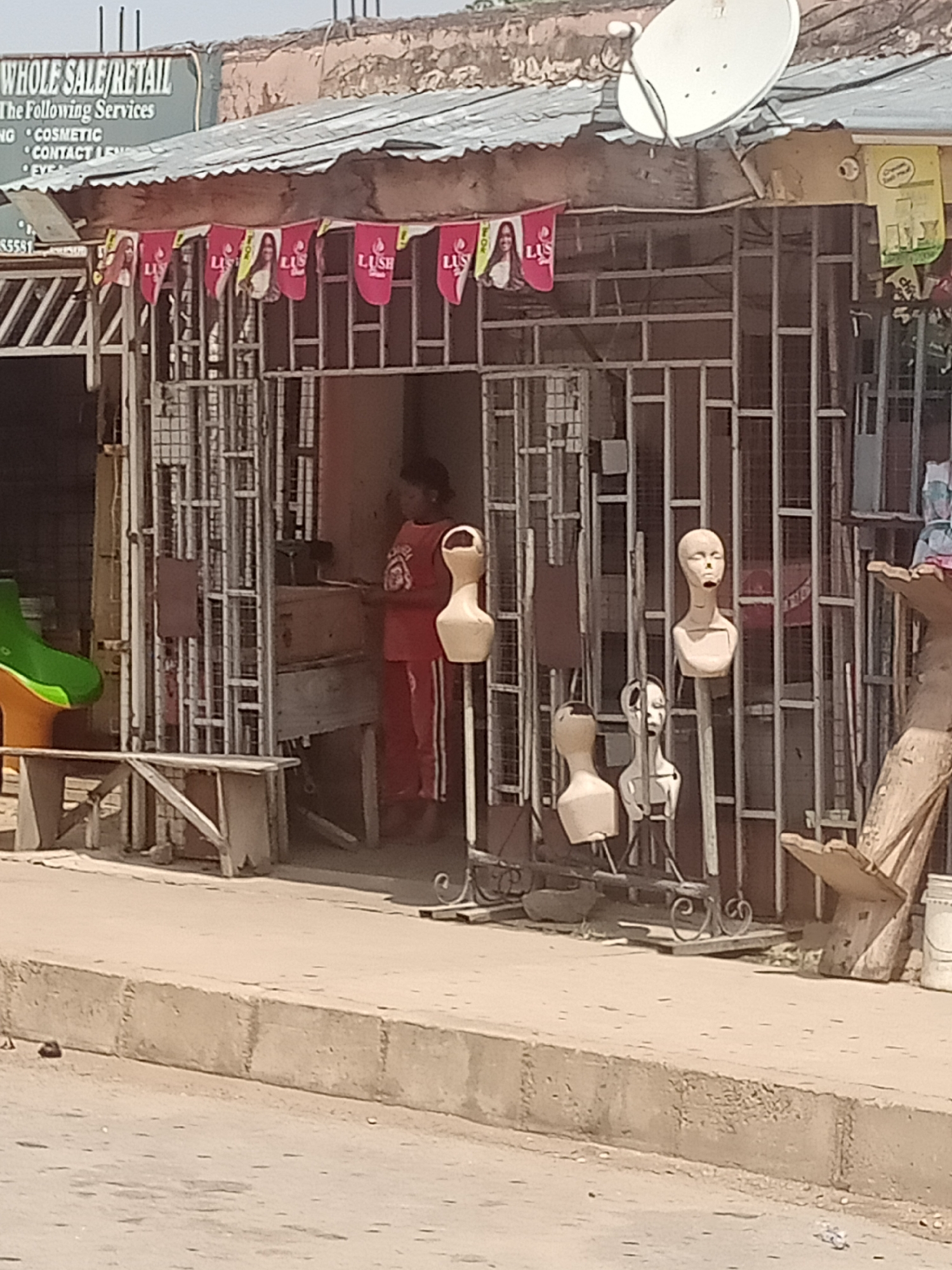 The image depicts the exterior of a retail establishment in Jalingo, Nigeria, situated at street level. The shop, likely a wholesale/retail business, features a corrugated metal roof and is secured by a metal grate. Banners bearing the brand "LUSH" and images of women are displayed above the entrance. Several mannequins, some with hair and others bare-headed, are positioned outside the shop, some on stands. A person in a red outfit is visible inside the shop, appearing to be working at a counter. In front of the shop, a wooden bench and a green and orange molded plastic object are present. The ground in front of the shop is paved with concrete, and a concrete curb separates it from a dirt road. A satellite dish is mounted on the roof. The overall scene suggests a commercial area in a developing urban environment.