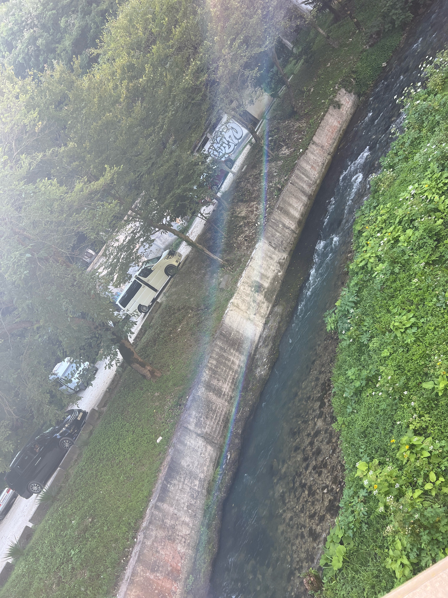 A body of water flows through a concrete channel with lush green vegetation on the right bank. On the left bank, a concrete wall is visible, separating the water from a grassy area with trees and parked cars. There is graffiti on the wall. A faint rainbow is visible in the upper left quadrant of the image, refracting sunlight. The scene appears to be an outdoor urban environment.