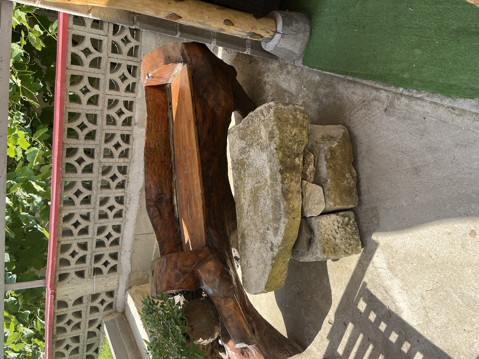 The image shows an outdoor seating area, likely a patio or garden space. In the foreground, a wooden bench crafted from natural, irregularly shaped wood is the central focus. The bench has a polished, reddish-brown finish. In front of the bench, a rustic coffee table is constructed from stacked, rough-hewn stones. A small potted plant with green foliage is visible on the left side of the bench, partially obscuring the lower portion of the wooden structure.
Behind the bench, a decorative concrete block wall with a repeating geometric pattern is present, topped with a red border. Green foliage, possibly grapevines, hangs from above the wall, suggesting a sunny outdoor environment. To the right of the bench, a thick, unfinished wooden post, likely supporting some overhead structure, stands on a concrete base. The ground surface is a mix of concrete and a patch of green artificial turf on the right. The scene is illuminated by bright sunlight, casting distinct shadows on the concrete. The presence of a decorative wall and the style of the seating suggest a private residence or a small hospitality venue. This scene is located in the city of Obzor, Bulgaria, likely in a backyard or garden area.