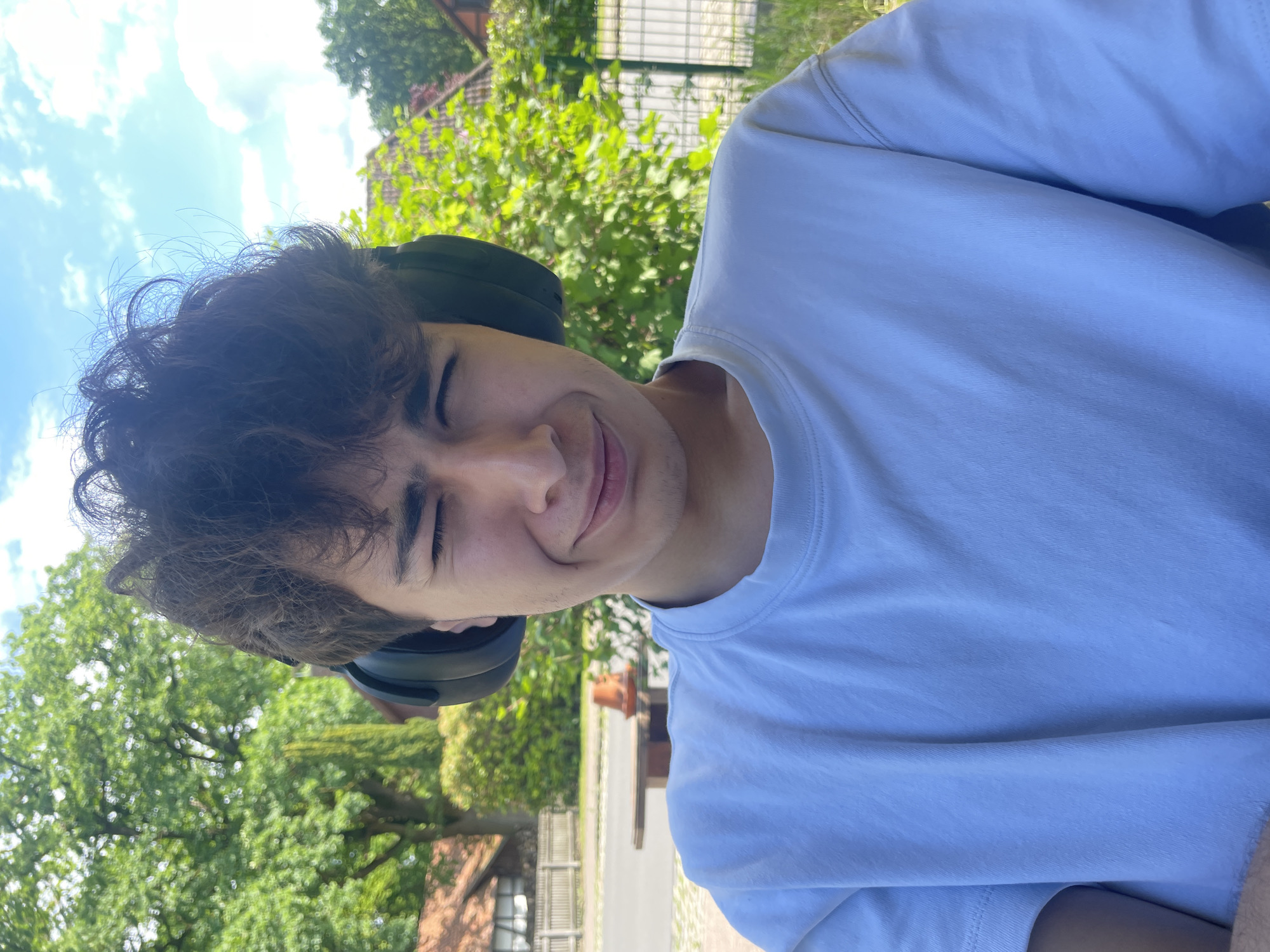 A young man with headphones is outdoors in Wienhausen, Germany. The man has dark, curly hair and is wearing a light-colored t-shirt. He is squinting his eyes and slightly smiling. The background includes trees, sky with clouds, and parts of buildings or structures. The photograph is taken at a tilted angle.