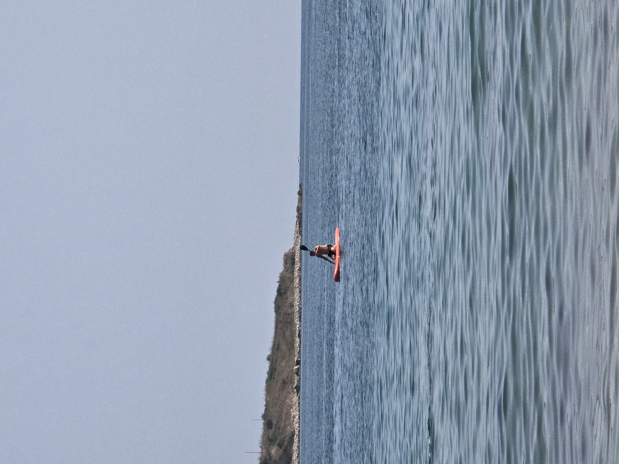 A person is paddleboarding on the calm, blue waters of the Black Sea in Sozopol, Bulgaria. The paddleboarder, wearing dark shorts, is seated and holding a paddle. In the background, a rocky, vegetation-covered shoreline stretches across the horizon, with a clear, pale blue sky overhead. The water is slightly rippled, and the overall scene conveys a sense of tranquility and outdoor recreation.
