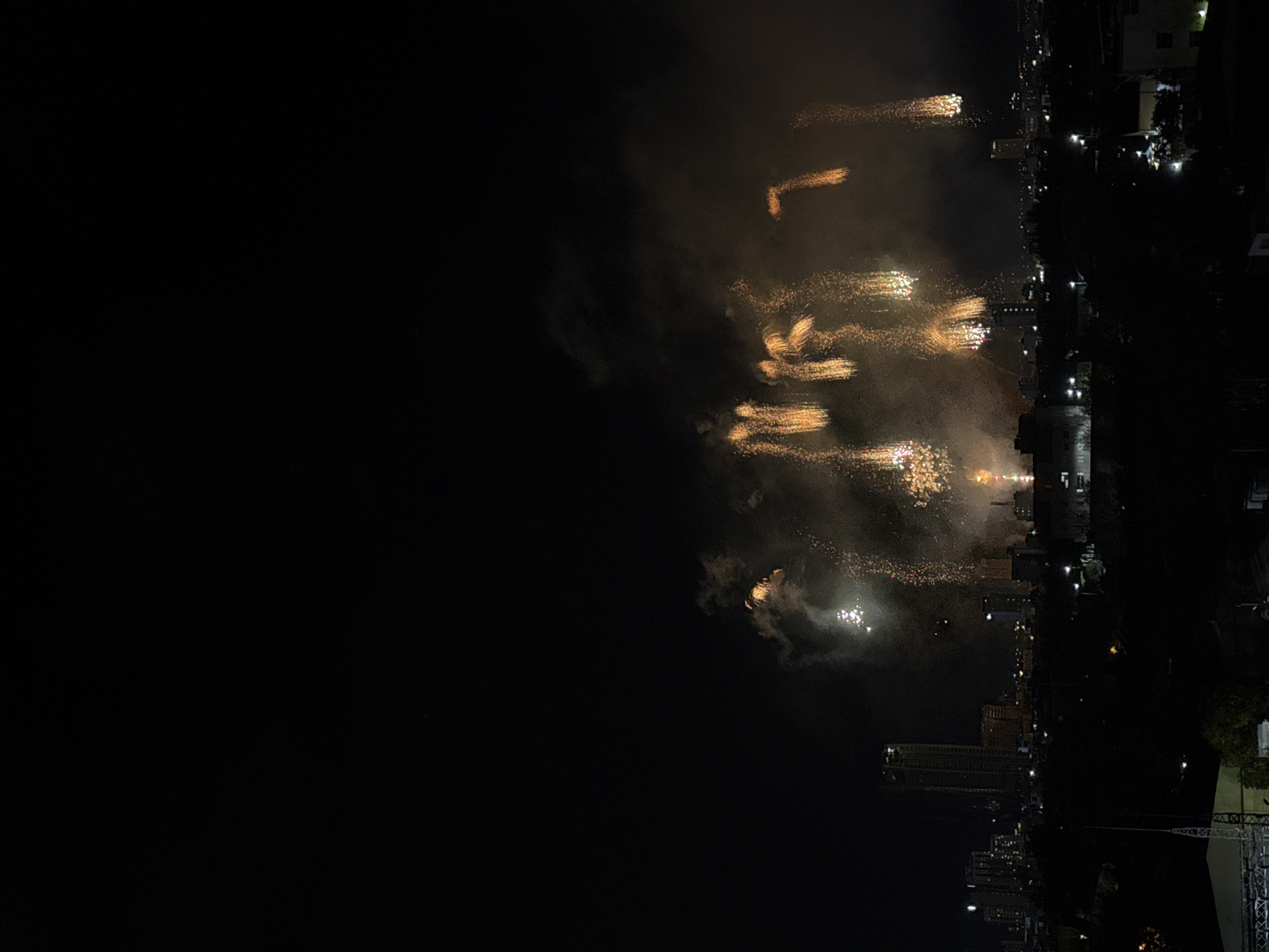 The image presents a nighttime panoramic view of a city, identified as Đà Nẵng, Vietnam, illuminated by a large fireworks display. The upper two-thirds of the frame consist of a dark, clear night sky. Below this, a dense cloud of greyish-brown smoke, heavily illuminated by the fireworks, dominates the mid-ground. Numerous golden-orange streaking fireworks are visible within this smoke, predominantly on the right side, creating a cascading or falling light effect. Brighter white bursts of fireworks are also interspersed among the orange trails, particularly towards the left-middle.
Beneath the fireworks and smoke, the cityscape extends across the lower portion of the image. On the left side of the skyline, a tall, slender high-rise building is prominently visible, its interior lights glowing. Various other urban structures of different heights are scattered throughout the mid-distance, marked by numerous points of artificial light from buildings and streetlamps. The immediate foreground at the bottom of the image is dark and features indistinct structures, including what appears to be part of a white roof or structure and some dark, grid-like elements on the far left. No individuals are discernibly visible in the scene.