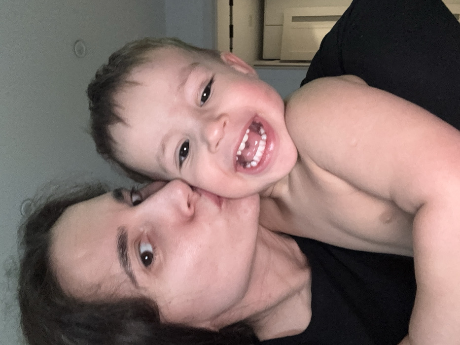An adult female subject with long dark hair, wearing a black top, is depicted kissing a shirtless child subject on the right cheek. The adult's visible left eye is directed towards the camera. The child, appearing to be a toddler with short dark hair, exhibits an open-mouthed smile, revealing a full set of primary teeth, and looks directly at the camera. The interaction occurs within an indistinct indoor environment. The background features a light-colored wall, a partially visible white door or cabinet on the far right, and a recessed ceiling light. The location is an indoor setting in the city of Chaikovsky, Russia.