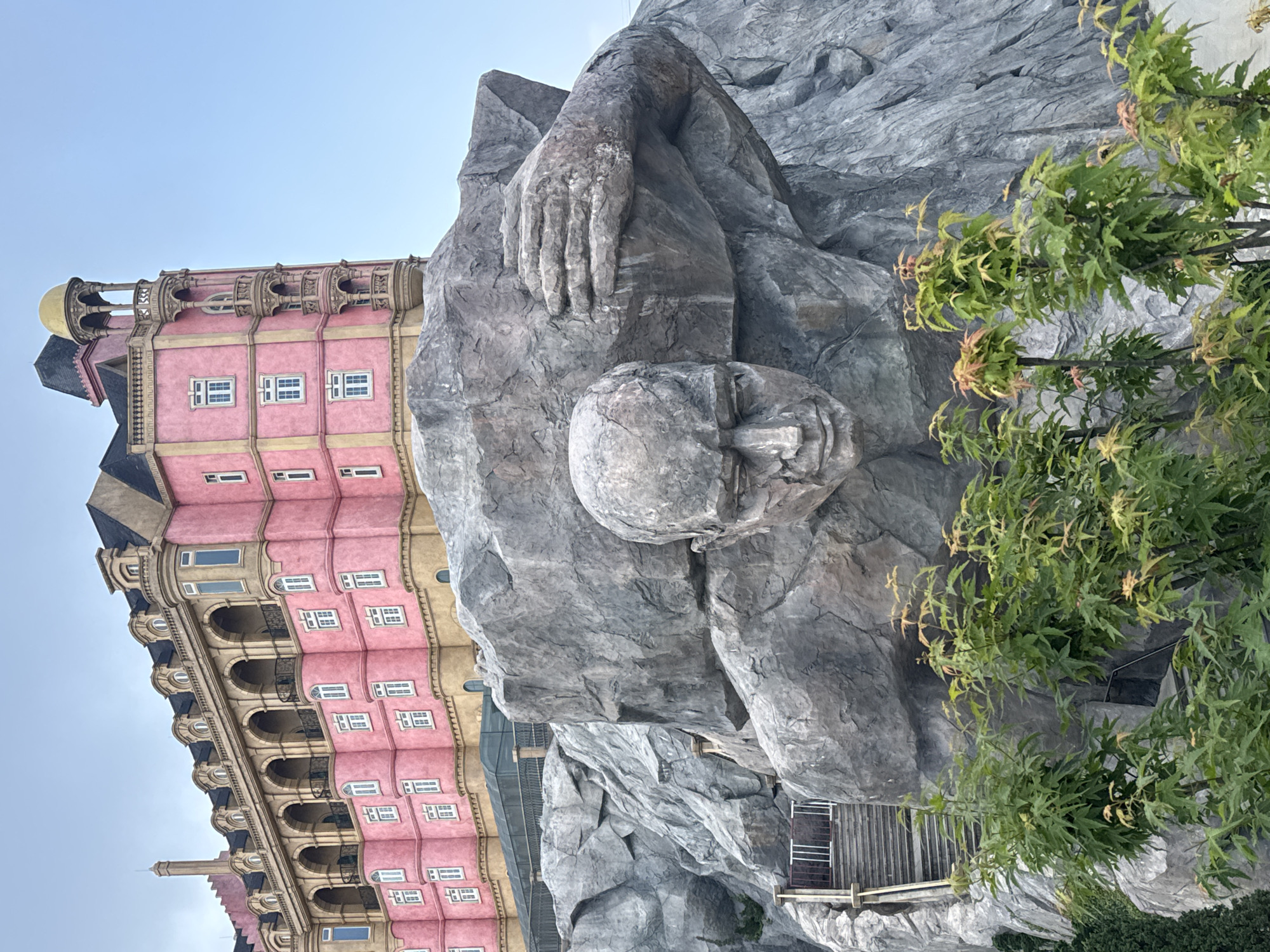 The image displays a unique architectural and sculptural scene, likely located in Sun World Ba Na Hills, Đà Nẵng, Vietnam. On the left, a prominent, multi-story building stands out with its vibrant pink or salmon-colored facade and intricate yellow-cream trim. It features numerous white-framed windows, elegant arched balconies with dark railings, and a multi-gabled roof adorned with dark tiles, golden domes, and ornate spires, evoking a classical European architectural style.
To the right, a colossal rock sculpture of a reclining human figure, carved from or integrated into a grey rock face, dominates the scene. The figure's head, with discernible facial features, is visible along with a large, textured hand resting on its body, conveying an impression of immense scale and strength. The rock's surface is rugged and detailed.
Green foliage, appearing as trees or large bushes with light green leaves, frames the right side of the rock sculpture, adding a natural element. A small wooden structure with railings is visible near the base of the rock formation on the far right. The sky overhead is a clear, light blue, suggesting fair weather during the daytime. The overall ambiance is one of grandeur and fantasy, created by the dramatic scale of the sculpture and the elaborate design of the building. No specific activity is depicted, and no text is visible within the frame.