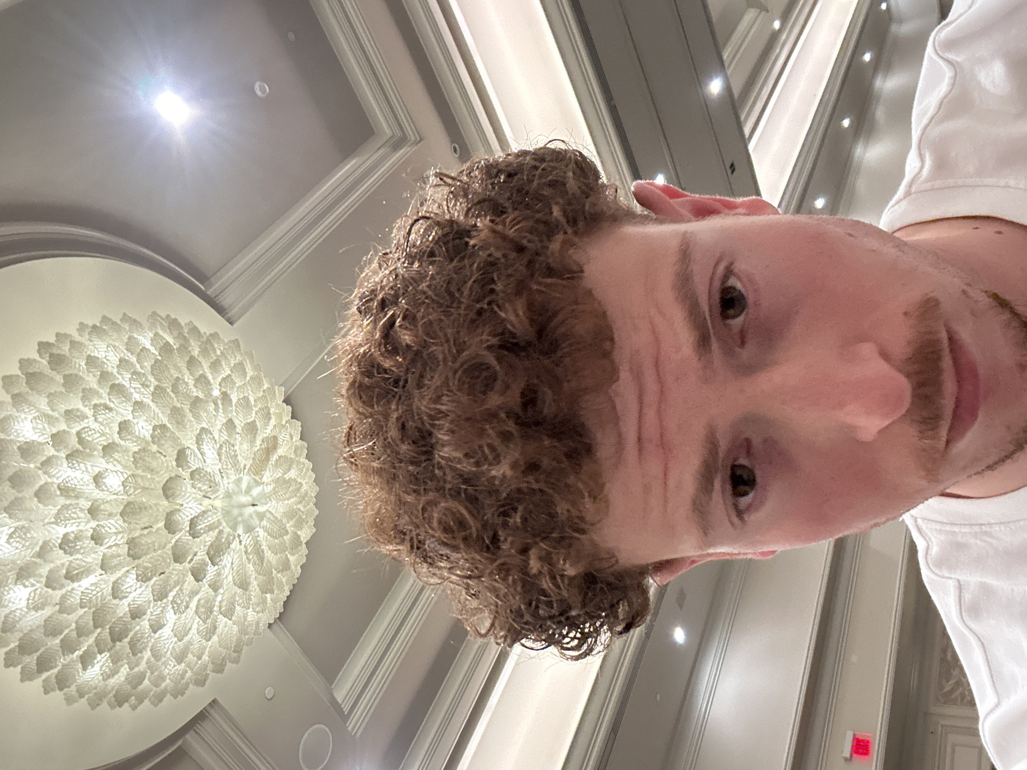 A low-angle, close-up shot features a person with curly brown hair, a light complexion, brown eyes, and a trimmed mustache. They are wearing a white shirt or top. The person's head is tilted slightly to the side. In the background, a large, decorative chandelier with a white, floral pattern is visible, suggesting an opulent interior setting. The ceiling appears to have recessed lighting and architectural moldings. The scene's location is likely in Aventura, United States of America.