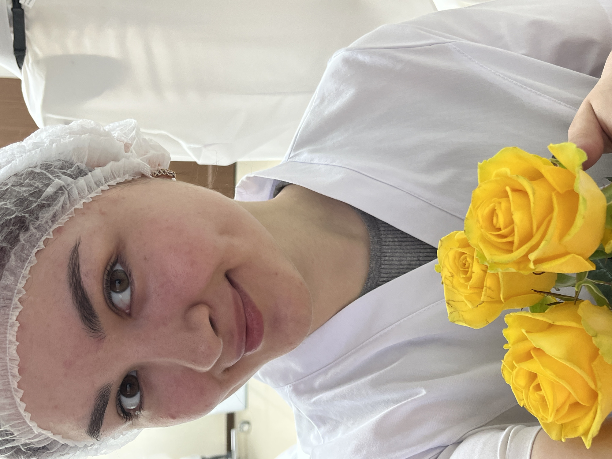 A young woman with brown eyes, wearing a white medical cap and a white coat, is holding a cluster of three yellow roses in her right hand. The background is predominantly white, possibly a clinical setting, with a glimpse of a wooden surface. The image is taken at an oblique angle. There is no verifiable information in the image that connects it to Neftekamsk, Russia.