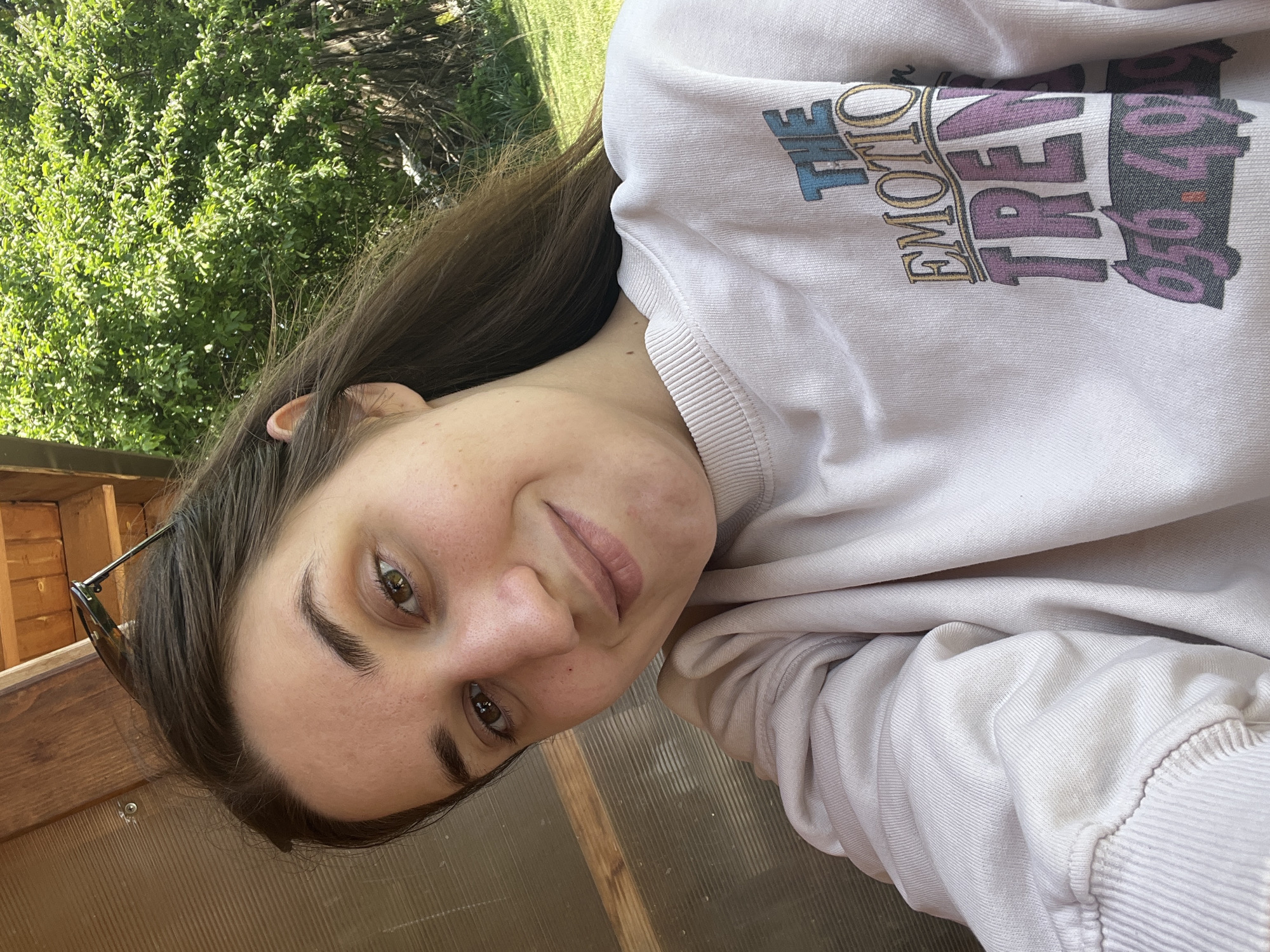 A young woman is photographed close-up, likely by herself, in Gorno Kamartsi, Bulgaria. She wears a light-colored sweatshirt with "THE EMOTIONAL TREND 6840" printed on it. The location appears to be partly enclosed, potentially a porch or greenhouse, with wooden framing visible. Lush green foliage is visible through an opening.