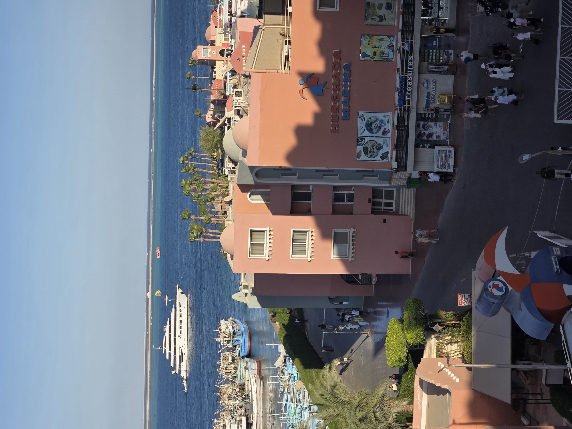The image depicts a view of Hurghada Marina in Hurghada, Egypt. In the foreground, there are several buildings with distinctive pink and teal facades, featuring arched windows and domed roofs. One building prominently displays the "HURGHADA MARINA" text and a colorful logo. Shops with glass storefronts are visible on the ground floor, showcasing various items like footwear, bags, and accessories. A paved walkway or street runs in front of these buildings, where several people are walking. Some are dressed in casual summer attire, while others appear to be tourists. In the background, a large white yacht is anchored in the blue waters of the Red Sea, alongside numerous smaller fishing boats. Beyond the marina, a sandy coastline is visible, with a clear blue sky overhead. Palm trees line the edge of the marina, adding to the tropical atmosphere. A paraglider can be seen in the distance, attached to a boat. The scene appears to be set during daylight, with the sun casting shadows on the buildings.