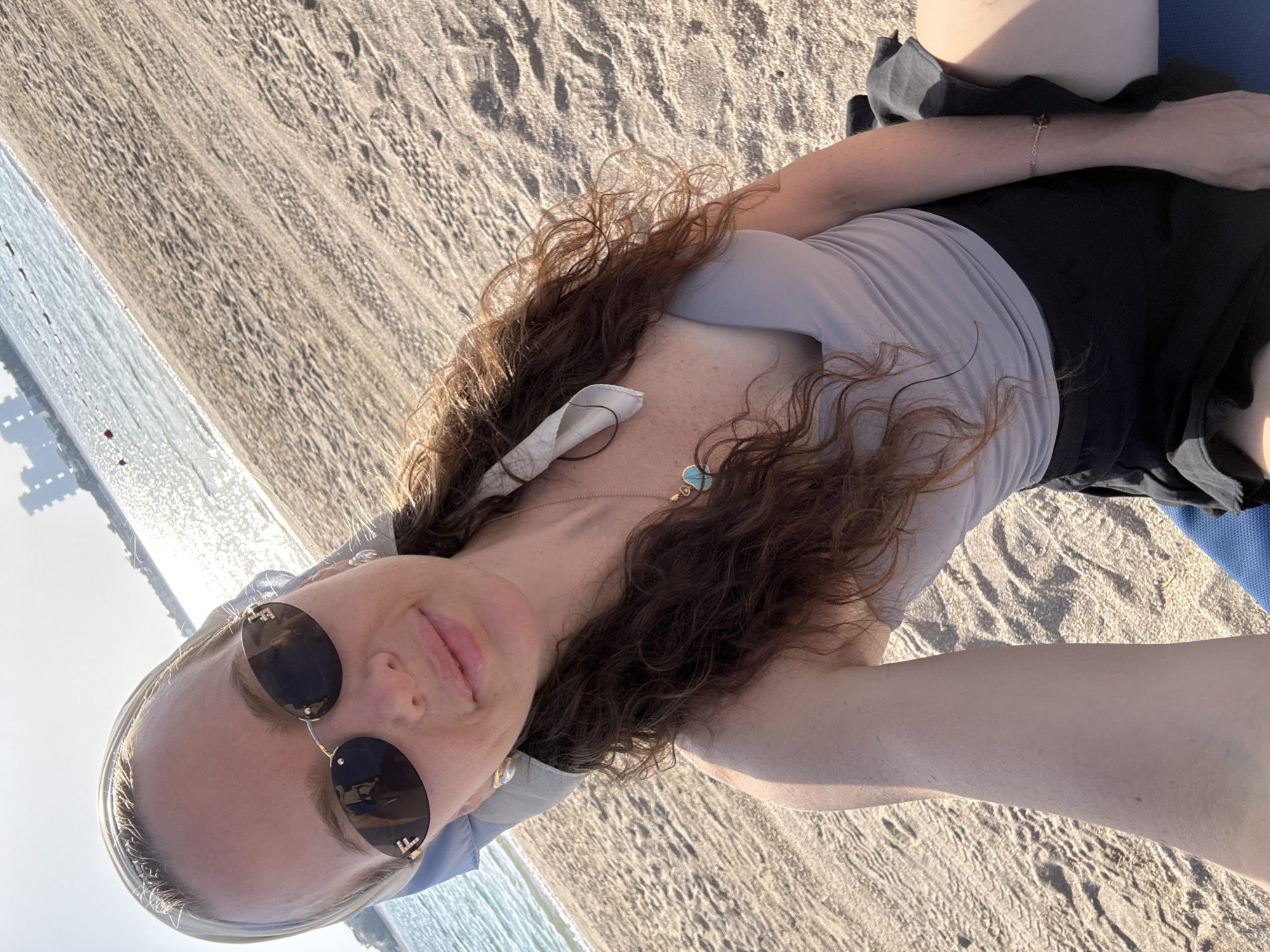 A woman is taking a selfie on a sandy beach with the ocean in the background. She is wearing sunglasses, a headscarf, and a grey tank top. The water is calm and reflects the sunlight. In the distance, buildings can be seen along the coastline. The sand is textured with footprints and light shadows. The sky is clear and bright, indicating a sunny day.