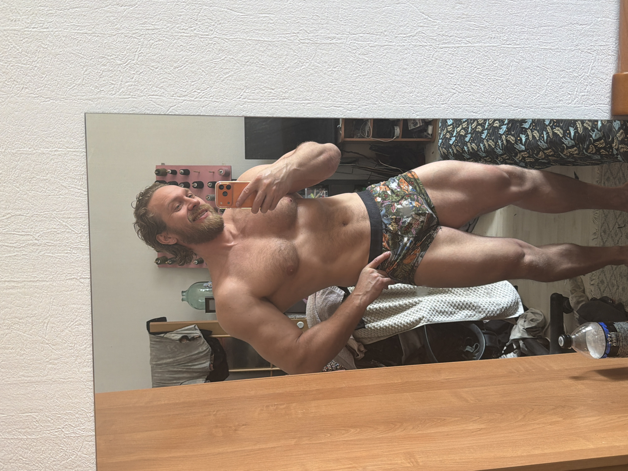 A muscular man with a beard and curly hair is captured in a mirror selfie, smiling broadly into an orange smartphone he holds. He is shirtless, showcasing his physique, and wears patterned boxer briefs. One hand is placed on his hip, while the other holds the phone to take the photo.
The setting appears to be an indoor living space, likely a bedroom or personal room, in Saint Petersburg, Russia, as evidenced by Cyrillic text visible on a water bottle. The reflection in the mirror reveals a somewhat cluttered environment behind the man, including a large glass carboy, a rack holding bottles (possibly wine), and various items of clothing and textiles, including a light-colored, textured blanket and patterned bedding. A clear plastic water bottle, partially visible on a wooden surface in the foreground, bears the brand name "EVIAN" and Cyrillic text such as "НАТУРАЛЬНАЯ МИНЕРАЛЬНАЯ ВОДА" (Natural Mineral Water) and "ПЕРЕРАБОТАННОГО ПЛАСТИКА" (recycled plastic).
The mirror is situated above a wooden surface, possibly a dresser or table. The wall outside the mirror's frame features a textured white wallpaper. The lighting suggests it is daytime. The man's cheerful expression conveys confidence and a positive mood.
