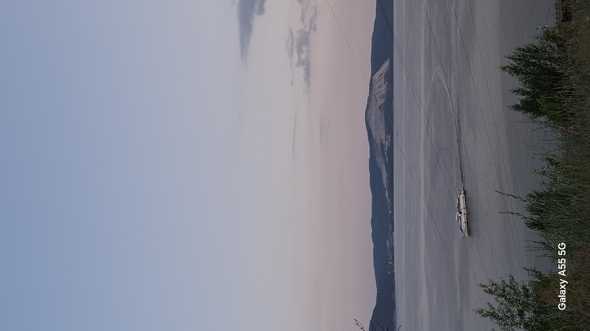 The image depicts a wide river or bay with a white passenger boat navigating the water. In the distance, a hazy, mountainous shoreline is visible, with a prominent white scar or deposit on one of the hills, possibly indicating industrial activity or a geological feature. The sky transitions from a pale blue at the top to a soft pink and purple hue towards the horizon, suggesting either dawn or dusk. Power lines are visible stretching across the frame, adding a man-made element to the natural landscape. The foreground is occupied by the dark silhouettes of trees and vegetation. The bottom left corner of the image contains a watermark that reads "Galaxy A55 5G". The scene is likely located on the Volga River in Tolyatti, Russia, where such landscapes and industrial markings on hills are common, particularly near the city's industrial zones or geological formations like the Zhiguli Mountains.