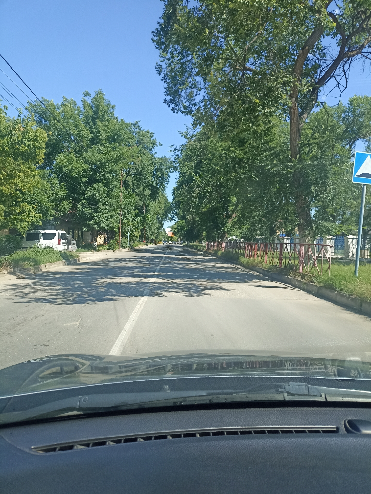 The image depicts a view from the interior of a vehicle, looking forward through the windshield. The vehicle appears to be driving on a two-lane asphalt road, with a white line dividing the lanes and a double white line on the right edge of the visible road. The road is flanked by trees on both sides, with a sidewalk and some buildings visible on the left. On the right side of the road, there is a red metal fence and a blue and white traffic sign indicating a speed bump. The sky is clear and blue. The scene is likely in Izberbash, Russia, given the context.