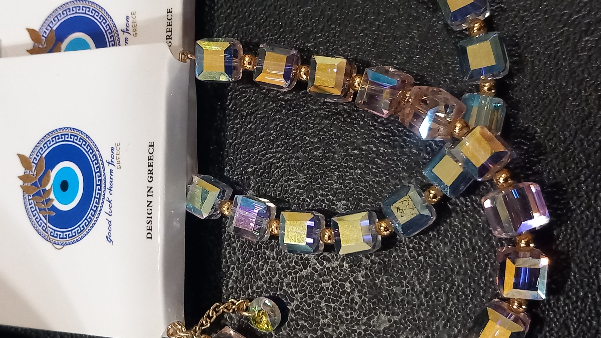 The image shows a close-up of a bracelet or necklace with square, faceted glass beads in iridescent shades of blue, purple, and yellow, interspersed with small gold beads. The bracelet is displayed on a black, textured surface. In the background, a white card with a blue and gold evil eye symbol is partially visible, along with text that reads "good luck charm from GREECE" and "DESIGN IN GREECE". This suggests the item is a souvenir from Greece, possibly purchased in the city of Kos, given the text. There are no people or discernible actions captured in the image.