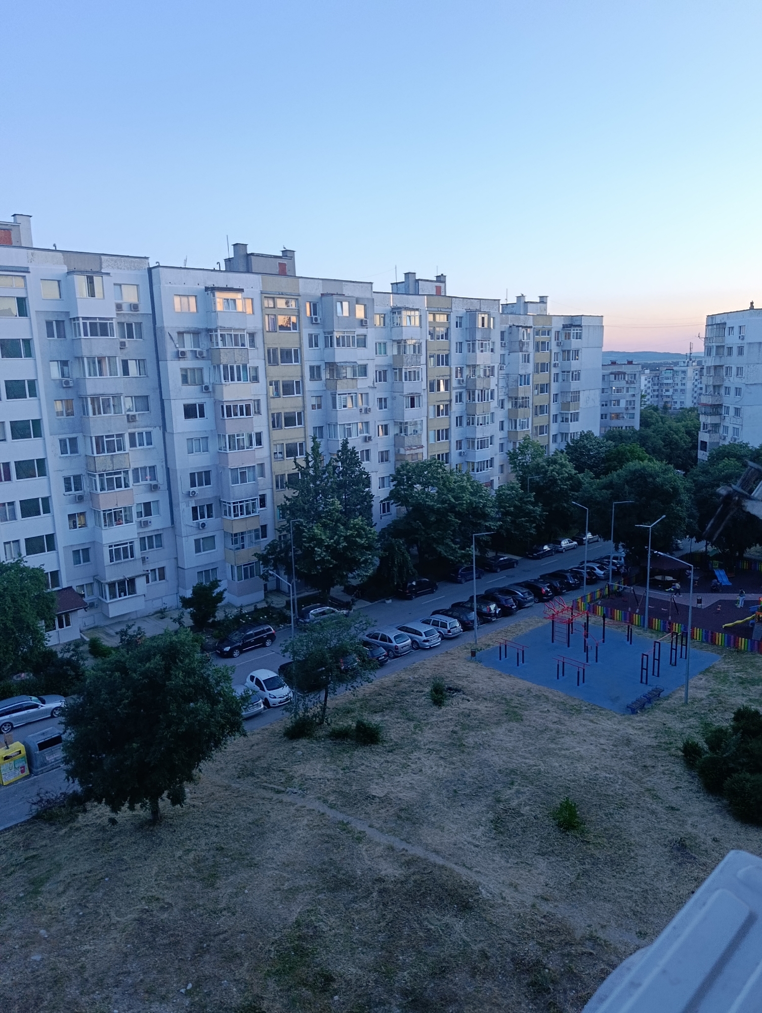 The image shows a view of a residential area in Varna, Bulgaria, at dusk. In the foreground, there is a patch of dry grass with some sparse vegetation. A large tree stands to the left, and a paved area with parking spaces is visible beyond it. Several cars are parked along the street. Further back, there are tall apartment buildings with numerous windows. A playground with colorful fencing and exercise equipment is situated in the middle ground. The sky is a clear, pale blue, transitioning to a soft pinkish hue near the horizon.