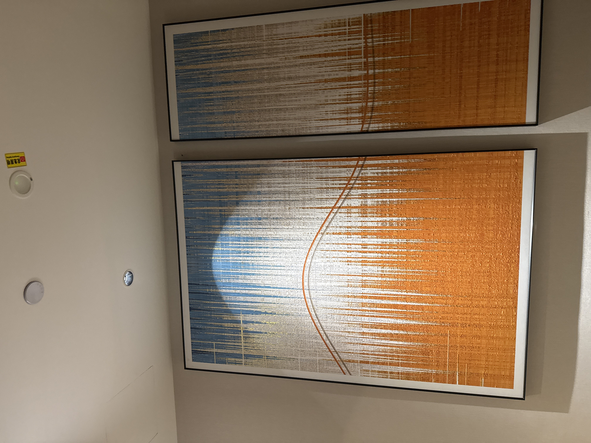 The image shows a modern interior with two large abstract paintings hanging on a neutral-colored wall. The paintings are framed in black and feature a textured design with vertical lines in shades of orange, blue, and gold, with a thin orange wavy line across the center of the left painting. Above the paintings, a white ceiling is visible, with two recessed lights and a circular smoke detector with a small yellow sticker attached. The sticker has Chinese characters on it. The overall scene suggests a contemporary and well-decorated space, likely in a commercial or residential building in Chaoyang District, China, given the presence of Chinese text on the smoke detector sticker.