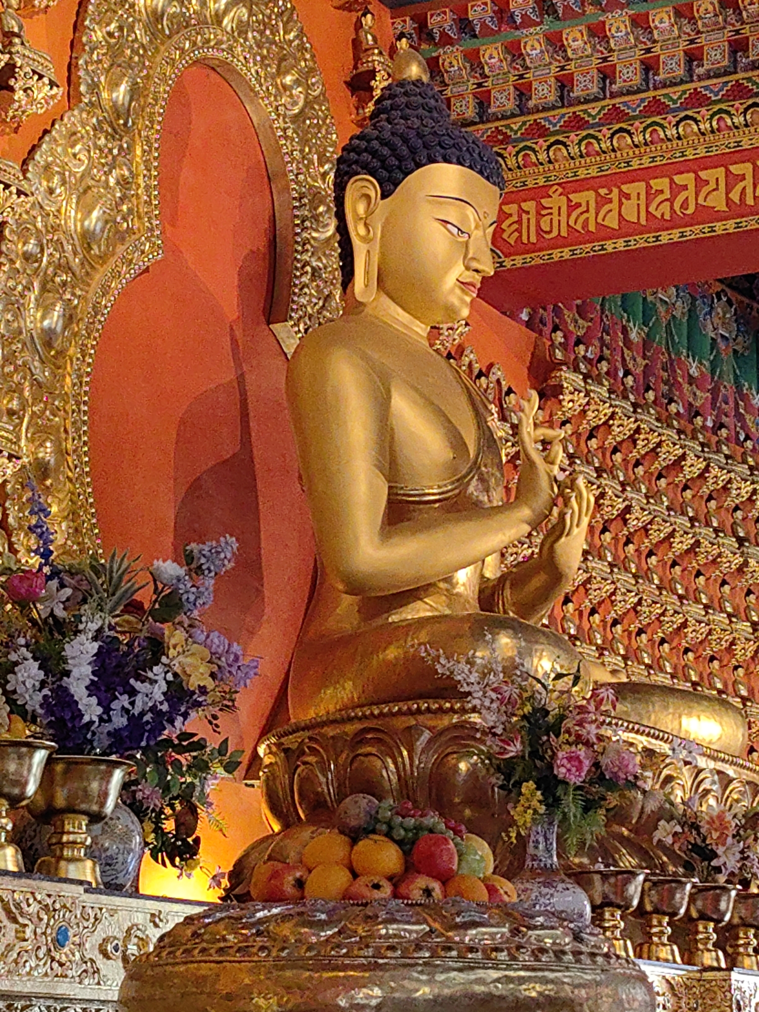 The image depicts a gilded statue of Buddha in a seated posture. The Buddha statue is adorned with intricate details and is positioned in front of an orange, arched backdrop. To the left of the statue, there is a golden, ornate frame surrounding the orange backdrop. The statue is decorated with offerings of flowers and fruits placed on a golden altar in front of it. Behind the statue, there are rows of smaller figures, possibly statues or paintings, and decorative elements on the ceiling and walls, suggesting an interior religious setting. The overall impression is one of reverence and opulence. This scene is located within the Boudhanath Stupa premises in Kathmandu, Nepal, a prominent Buddhist pilgrimage site.