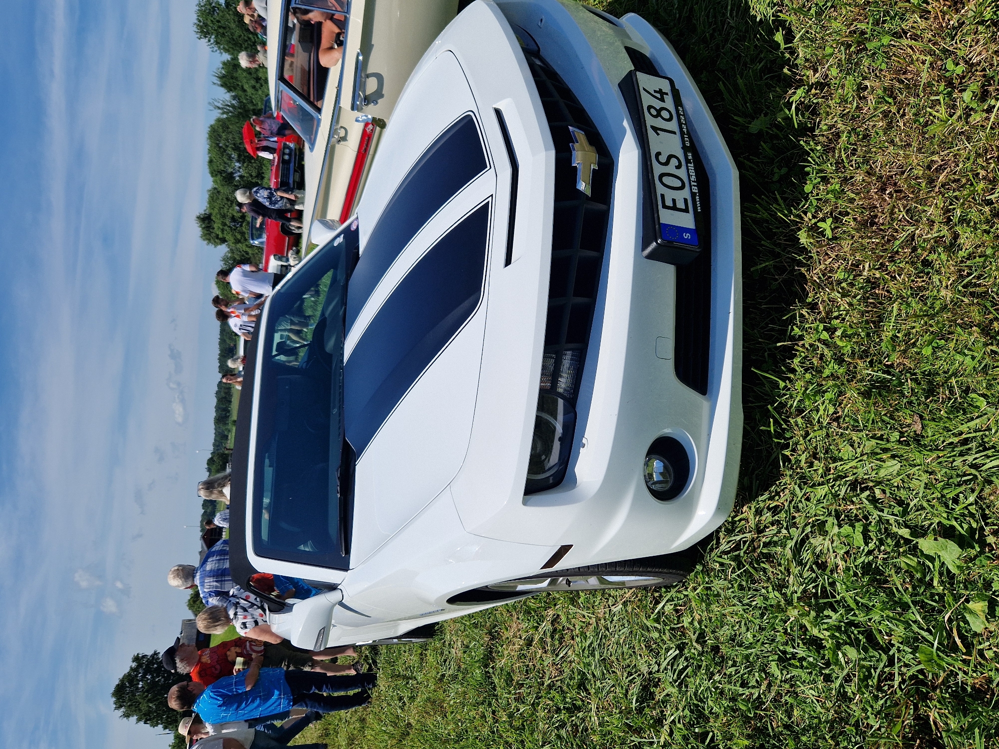 This image depicts a white Chevrolet Camaro convertible parked on a grassy field under a clear blue sky with scattered clouds. A group of people are gathered around the car and in the background, some appearing to be attending an outdoor event. The car has black racing stripes on its hood. A license plate reading "EOS 184" is visible on the front of the car, along with a website and phone number. The car is the primary subject, positioned in the foreground and angled slightly towards the viewer. The environment appears to be a rural or park-like setting. The scene is consistent with a car show or community gathering in Edenberga, Sweden, indicated by the Swedish license plate and the overall ambiance.