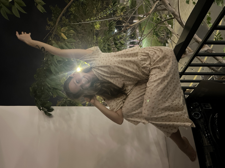 A woman in a floral nightgown poses outside at night. She leans back, with one arm raised and the other near her face. Lush greenery is above and behind her, lit by a bright light. She is barefoot with one foot visible. A modern staircase with black metal railings is visible on the right. Below her is a white wall.