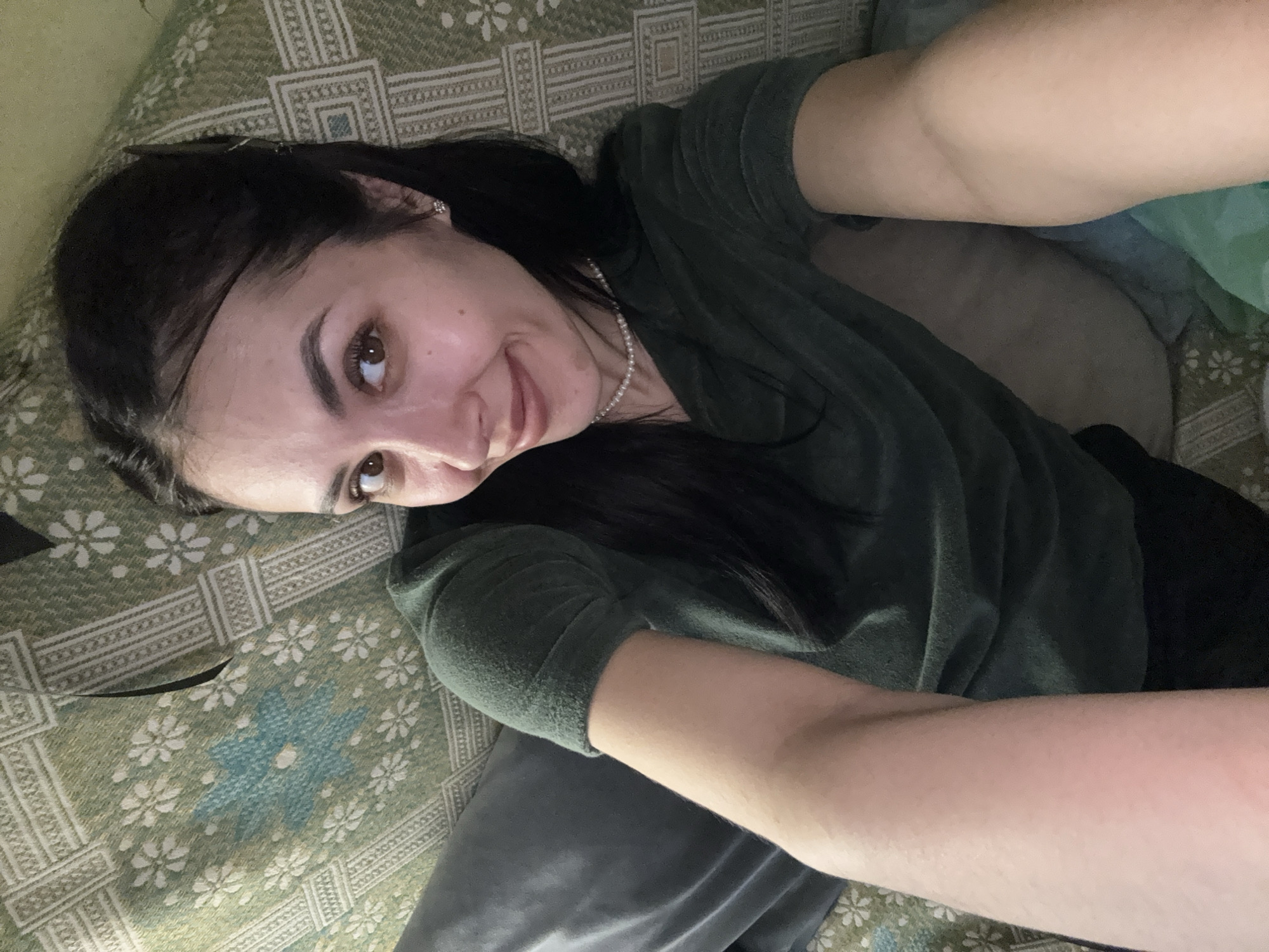 A young woman is captured in a selfie-style photograph, lying down in an indoor setting in Votkinsk, Russia. She is positioned diagonally across the frame, looking directly towards the camera with a gentle, relaxed smile.
The woman has dark hair, styled down, and expressive dark brown eyes. She is wearing a dark green, possibly fleece or textured, long-sleeved top, complemented by a delicate white pearl necklace. Her skin tone is fair. Her left arm is extended towards the camera, indicating she is holding the device for the photograph, while her right arm is bent. Parts of dark pants or leggings are visible below her top.
She is resting on a patterned bed or couch cover. The fabric is predominantly green, adorned with white floral designs and distinct blue floral patterns in the center, all framed by intricate geometric borders. The overall impression is one of comfort and ease. No visible text is present in the image. The lighting suggests an indoor environment, though the specific time of day is not discernible.