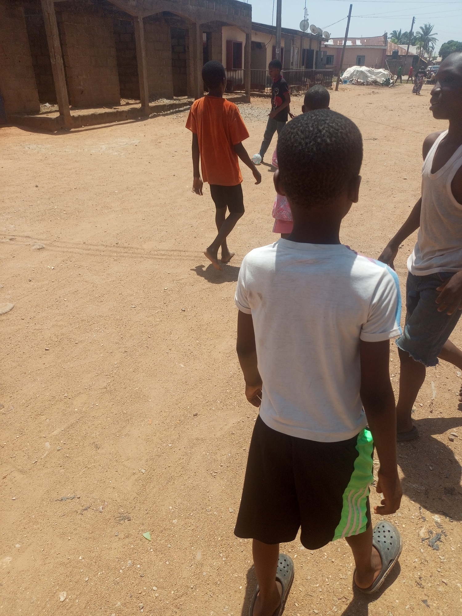 Several children stand on a dirt road in Sabon Tasha, Nigeria. One child wears an orange shirt and dark shorts, walking barefoot. Another child, wearing a white t-shirt with blue stripes on the sleeves and dark shorts with green accents, faces away from the camera. A third child wears a white tank top and denim shorts. Buildings are visible in the background, with satellite dishes on some rooftops.