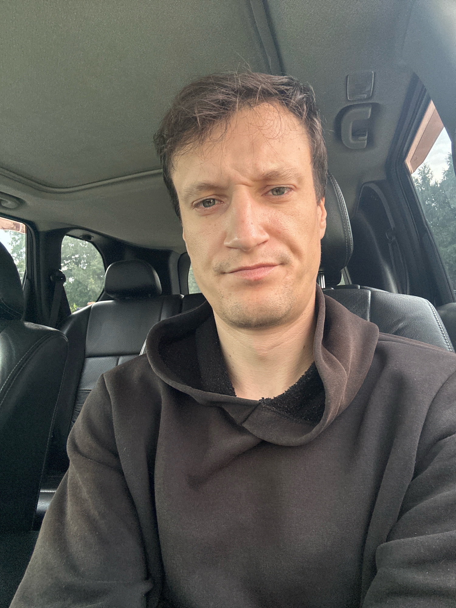 The image shows a medium shot of a man sitting in the driver's seat of a car. He is looking directly at the camera with a slightly furrowed brow and a neutral expression. He is wearing a dark brown hooded sweatshirt. The interior of the car is visible, including black leather seats and the headliner with a sunroof. Outside the car, trees and greenery can be seen through the windows. The man is positioned in the foreground, and the background is slightly blurred. The location is specified as Makiv, Ukraine, though there are no specific visual cues in the image to confirm this location.