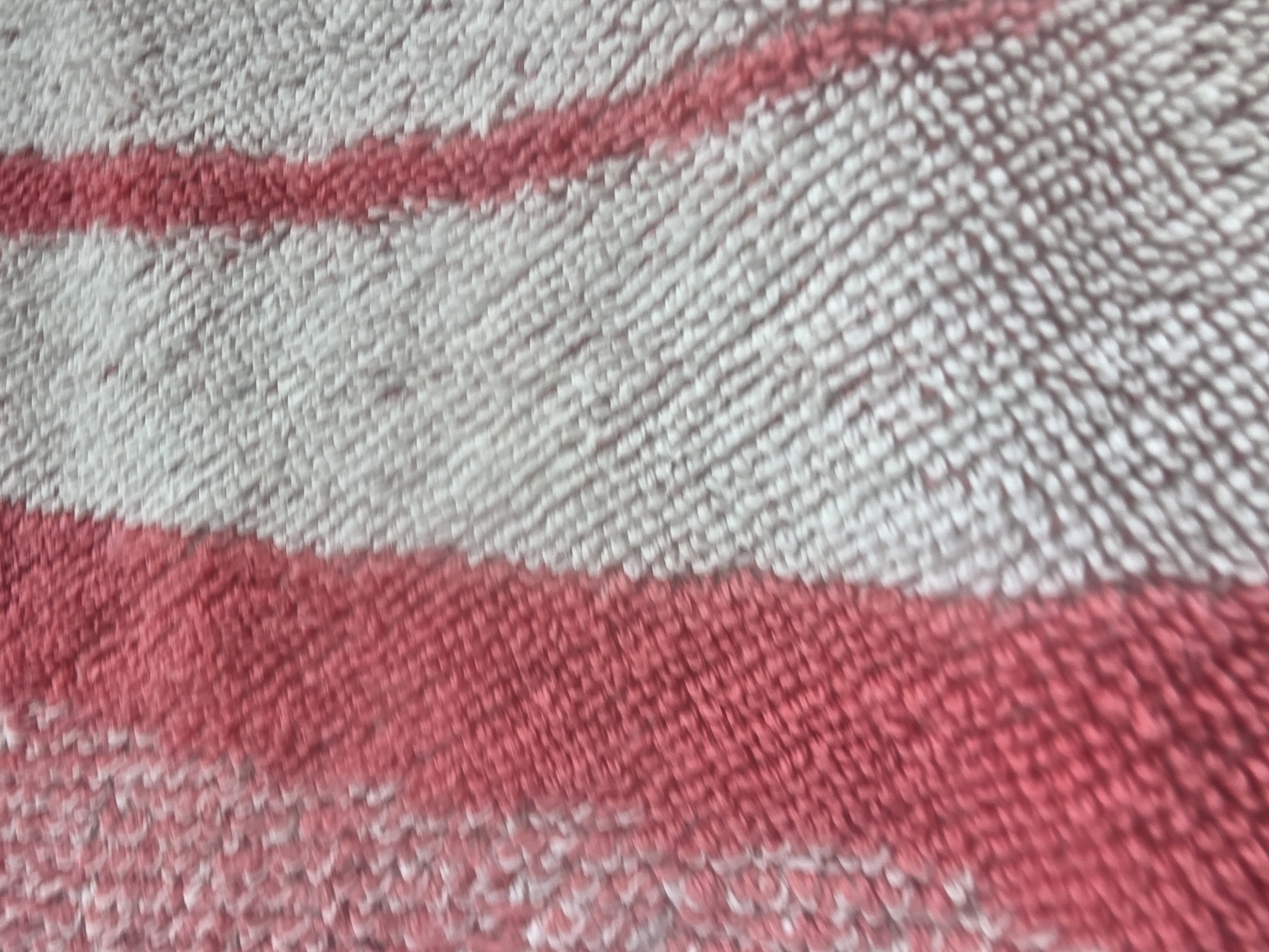 The image shows a close-up, blurry view of a textured surface with alternating stripes of white and pink. The texture appears to be that of a terry cloth or woven fabric. The stripes run horizontally across the frame, with one pink stripe at the top, followed by a white stripe, then another pink stripe, and finally a predominantly white section at the bottom. Due to the blurriness and lack of context, it is not possible to determine the specific location in the city of Dubno, Ukraine, nor are there any discernable people, actions, or interactions visible. The scene is simply a detailed but indistinct texture.
