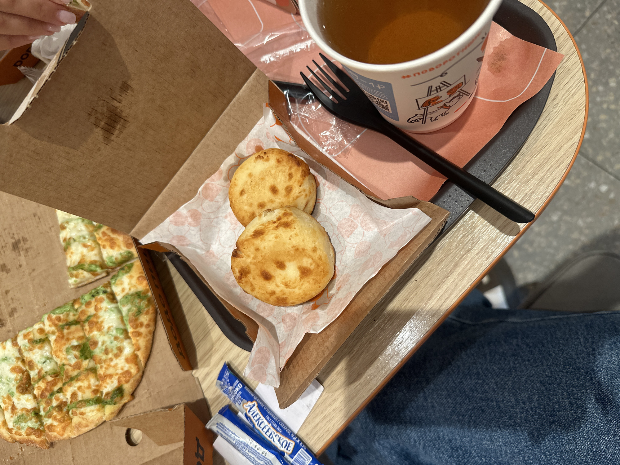 The image shows a table in a food establishment, likely a fast-food restaurant, in the city of Sverdlovskiy, Russia. On the table, there is a pizza with green sauce and cheese, cut into squares, in a cardboard box. Next to the pizza are two small, round, golden-brown baked goods, possibly buns or rolls, served on a piece of patterned paper. There are also two small blue packets, likely condiments or seasonings, and a black plastic fork. In the foreground, a white paper cup filled with a light brown liquid, possibly tea, sits on an orange paper napkin. The table surface is made of light-colored wood. The overall scene suggests a casual meal setting.