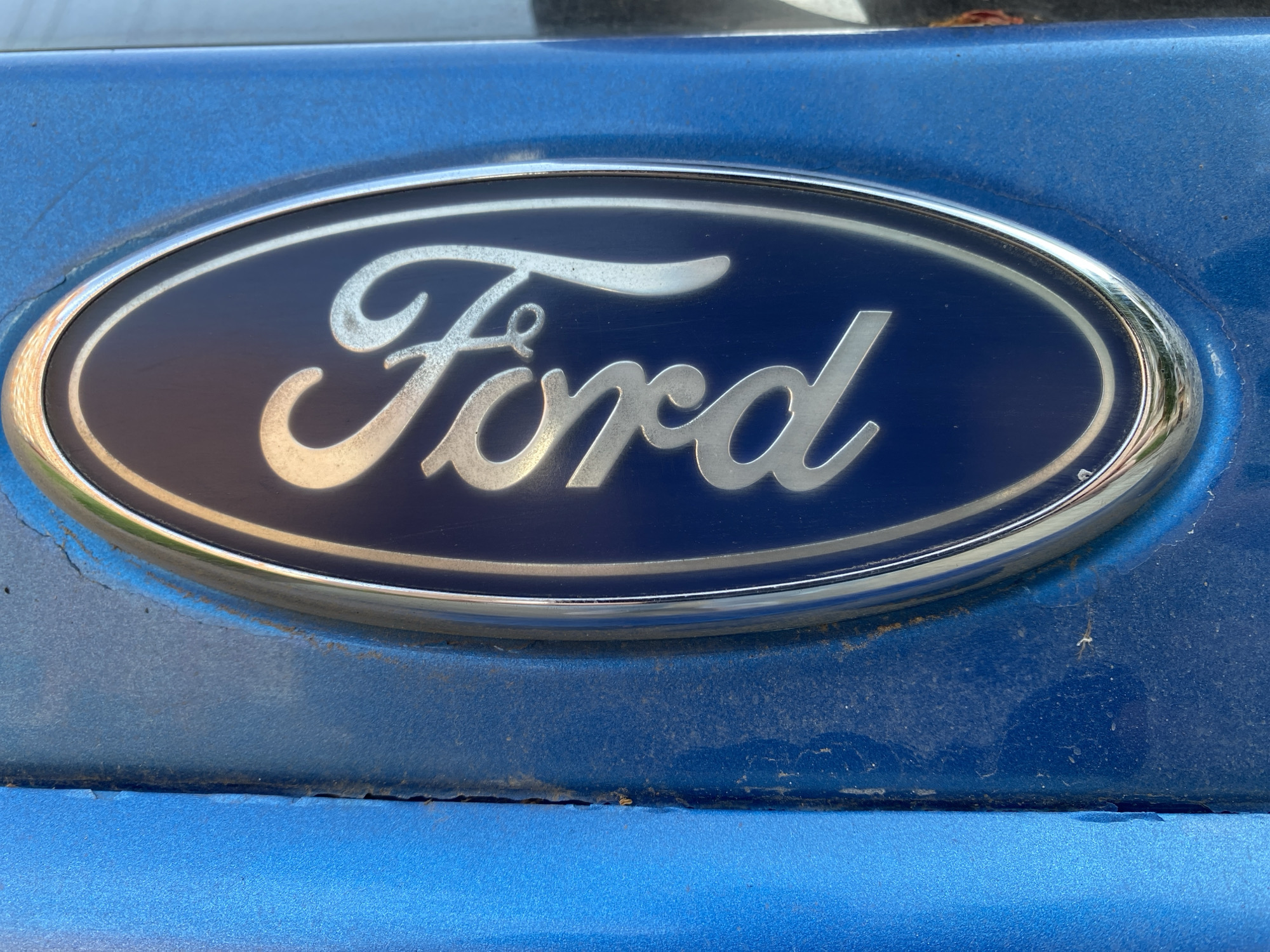 The image is a close-up, low-angle shot of a blue Ford emblem on a blue car. The emblem is oval-shaped with a dark blue background and the word "Ford" written in silver. The car's paint is chipped and weathered, particularly around the emblem, showing signs of rust. The background is out of focus, but appears to be outdoors with natural light. The image does not contain any discernible elements that would indicate its specific location within the city of Chaikovsky, Russia.