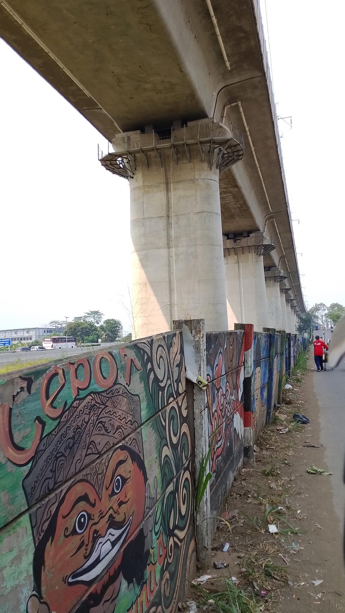 An eye-level, medium shot captures a mural on a wooden fence beneath an elevated concrete structure, likely an overpass, in Bojongsoang, Indonesia. The mural features a stylized face with large eyes and a wide smile, adorned with a traditional patterned headdress. The wall is comprised of horizontal wooden planks painted with various vibrant colors and designs. To the right of the mural, a paved road runs alongside, with a person in a red shirt walking away from the camera. The environment is somewhat unkempt, with debris visible on the ground near the wall and road. In the background, traffic can be seen on a highway, with buildings and trees lining the horizon. The sky is bright and appears to be daytime, with the sun casting shadows from the concrete structure.