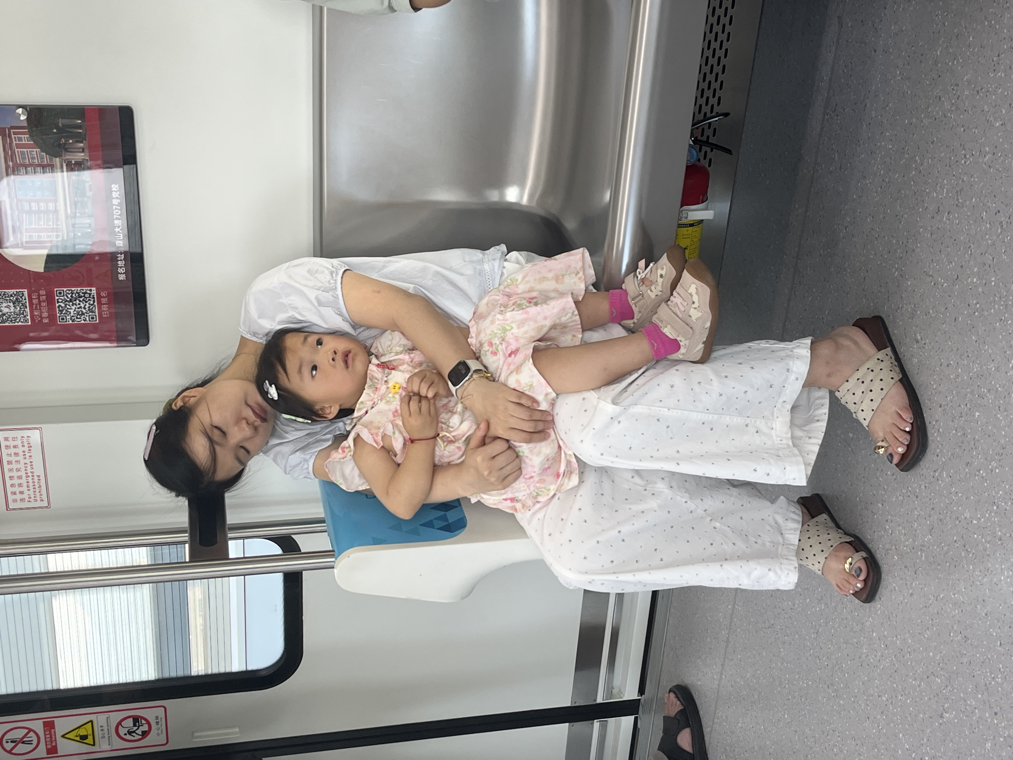 The image shows a woman and a young child on a train, likely in Tiaodeng, China, based on the presence of Chinese text on signage. The woman, with her eyes closed, is holding the child on her lap. The child, wearing a pink floral dress and pink socks, appears awake and is looking to the side. They are seated next to a metal railing and a blue and white seat. The train interior features light-colored flooring and walls, with windows showing an outdoor view. There are various signs in Chinese on the train's interior, one of which indicates the location as "Tiaodeng Avenue 707" in Chinese characters. The overall scene depicts a quiet moment of rest during a train journey.