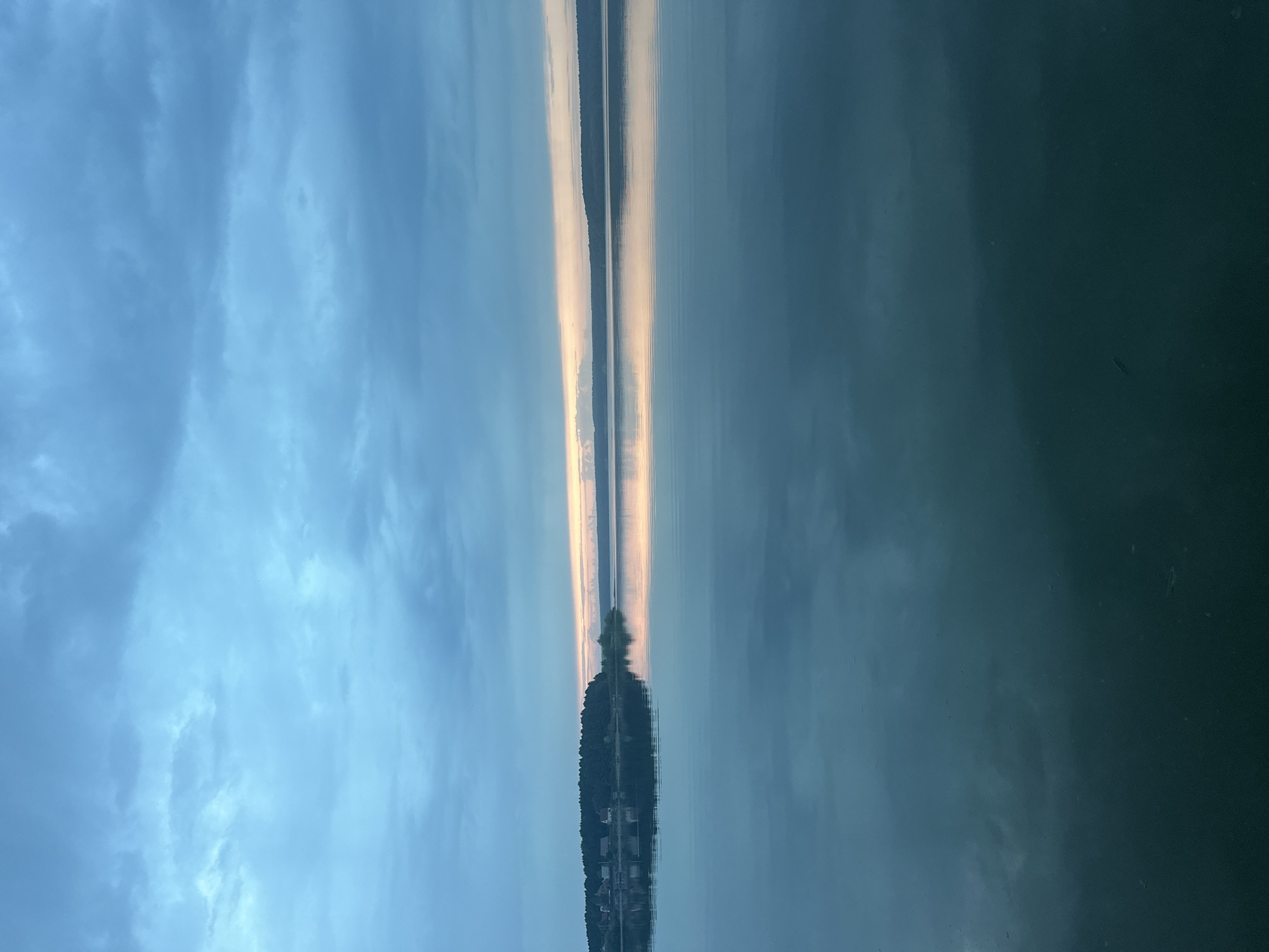 The image depicts a still body of water, likely a lake, with reflections of the sky and land. The sky is a mix of light blue with some clouds. A stretch of land, possibly a peninsula or an island, is reflected in the water. There are indications of trees and possibly buildings on the land. The water is a darker shade of blue-green, suggesting depth or different lighting conditions. The vertical orientation of the photo has been altered. The location of Aviator, Russia has been implied.