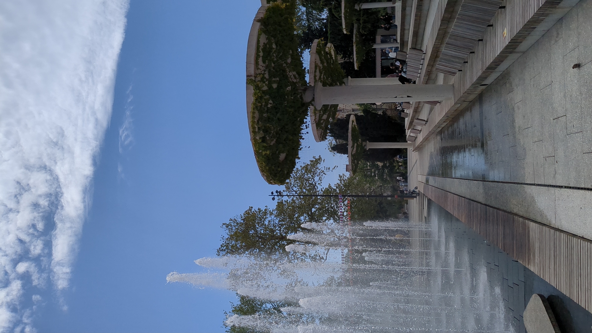 The image shows a fountain in the city of Yalta, Ukraine. The fountain's jets of water create a dynamic display against the blue sky. Above the fountain, trees provide a backdrop to a pergola-like structure adorned with green vines. People can be seen sitting on benches near the structure. A paved walkway, reflecting the wetness from the fountain, stretches alongside the fountain. The sky contains both blue areas and fluffy clouds.