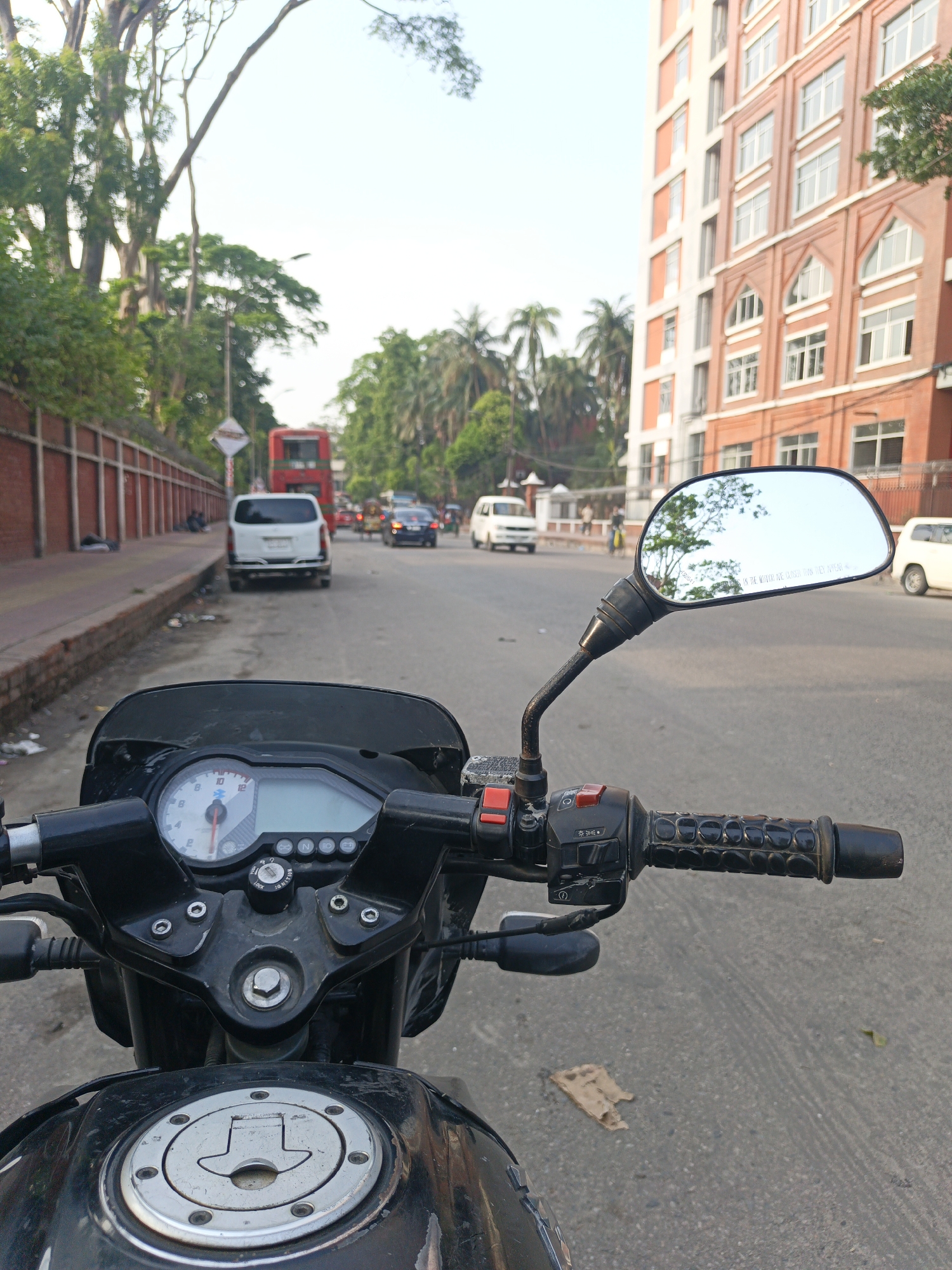 The image, taken from a motorcycle rider's point of view, depicts a street scene in Unknown, Unknown. The foreground features the motorcycle's handlebars, instrument panel, and gas tank. The street is visible ahead, with various vehicles, including a red double-decker bus, a white van, and other cars. Buildings line both sides of the street, with a prominent red brick building on the right. Trees and greenery are interspersed along the street and behind the buildings. Debris litters the roadside, and what appear to be individuals are laying on the side of the road. The sky is overcast.