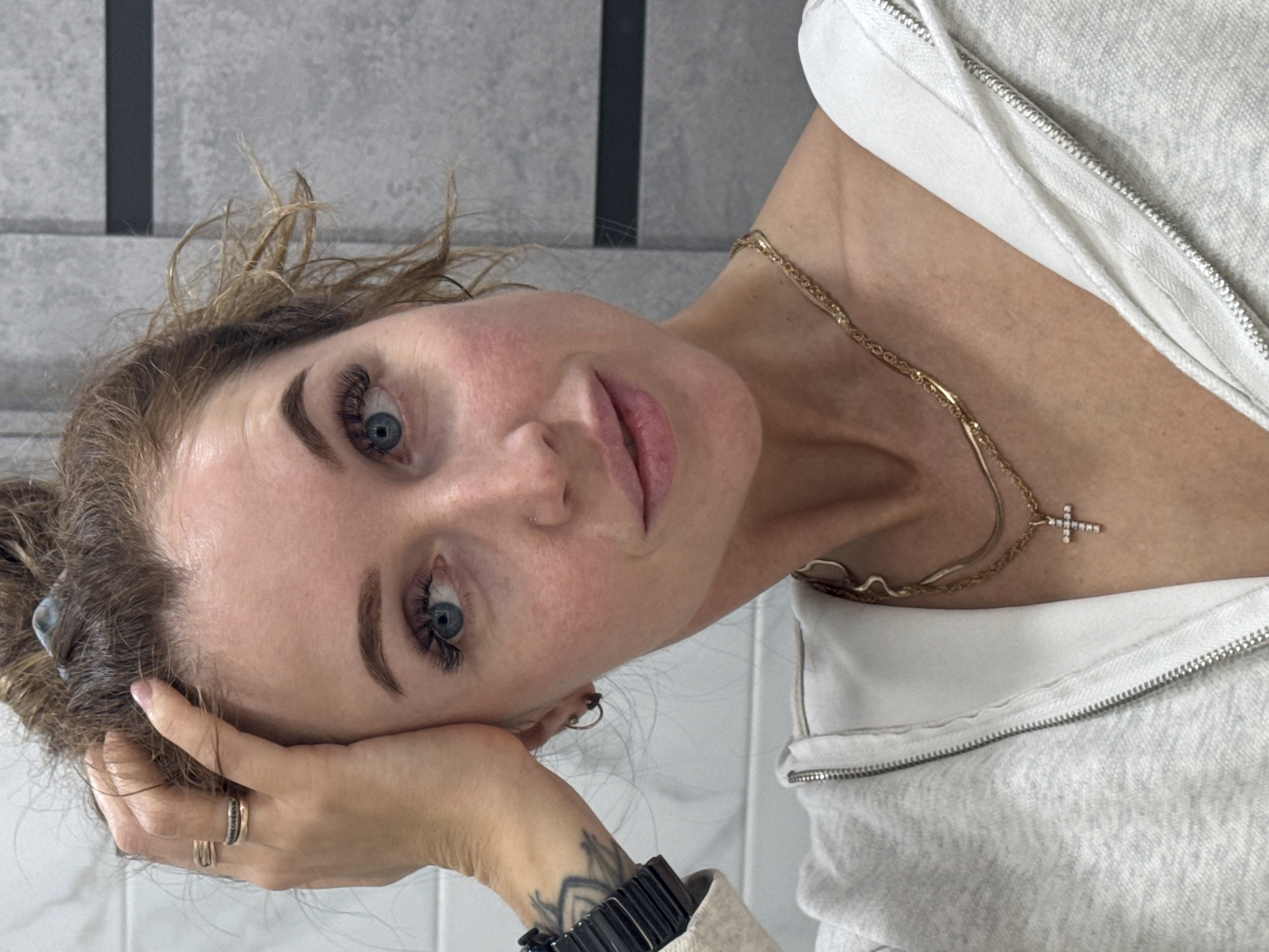 A close-up portrait shows a woman with her head tilted, looking directly at the camera. She has light brown or blonde messy hair, blue eyes with prominent eyelashes, and defined eyebrows. Her lips are full and slightly parted.
She is wearing a light grey or off-white zippered hoodie. Around her neck are two gold-colored necklaces, one featuring a small, sparkling cross pendant. Her left hand, adorned with a gold ring and a dark-colored ring, rests near her temple. A dark watch or bracelet is visible on her right wrist, along with a partially visible geometric tattoo. She has a small hoop piercing in her right nostril and a stud piercing above it.
The background consists of light grey, textured walls with a vertical dark stripe, suggesting a modern interior space, possibly a bathroom, in Chaikovsky, Russia. A plain white tiled surface is also visible in the lower left portion of the image. The woman's expression is calm and direct. No text is visible in the image.
