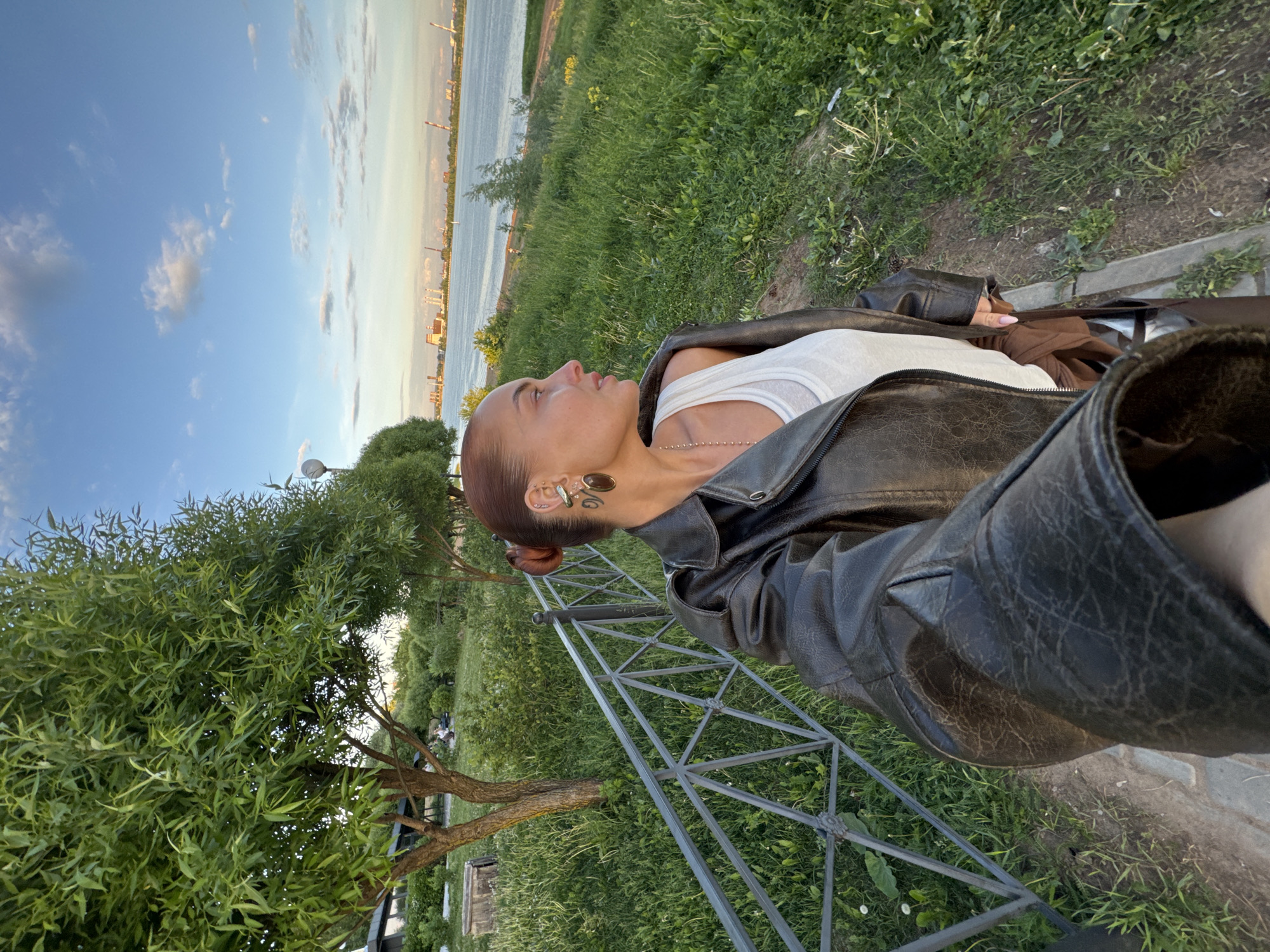 This image, taken in Izhevsk, Russia, captures an outdoor scene featuring a person in a verdant park-like setting beside a body of water. The photo is taken at eye-level with the subject, from a slightly lower angle.
The primary subject is a person, visible from the chest up, positioned on the left side of the frame. They have light brown or auburn hair pulled back into a neat bun. They are wearing a white tank top layered under a dark, textured leather-look jacket. The person's head is slightly tilted back, and they are looking towards the right, seemingly observing the distant view with a calm and contemplative expression. Notable details include a beaded necklace, and on their visible left ear, multiple earrings including a cuff, a stud, and a large oval or teardrop-shaped brown earring. Small, intricate designs resembling temporary tattoos are visible near their temple/ear.
The background features a wide body of water, possibly a river or lake, stretching horizontally across the mid-ground. On the far bank, a distant city or industrial skyline is visible under a clear sky dotted with scattered clouds, indicating late afternoon or early evening light. The sun casts a soft, warm glow. The foreground and mid-ground are dominated by lush green grass. On the left, a leafy tree with slender, willow-like leaves is prominent. To the right of the person, a dark metal lattice structure, possibly part of a fence, railing, or support, extends from the foreground into the mid-ground. A narrow concrete or paved edge runs along the grassy area in the lower right. No visible text is present in the image.