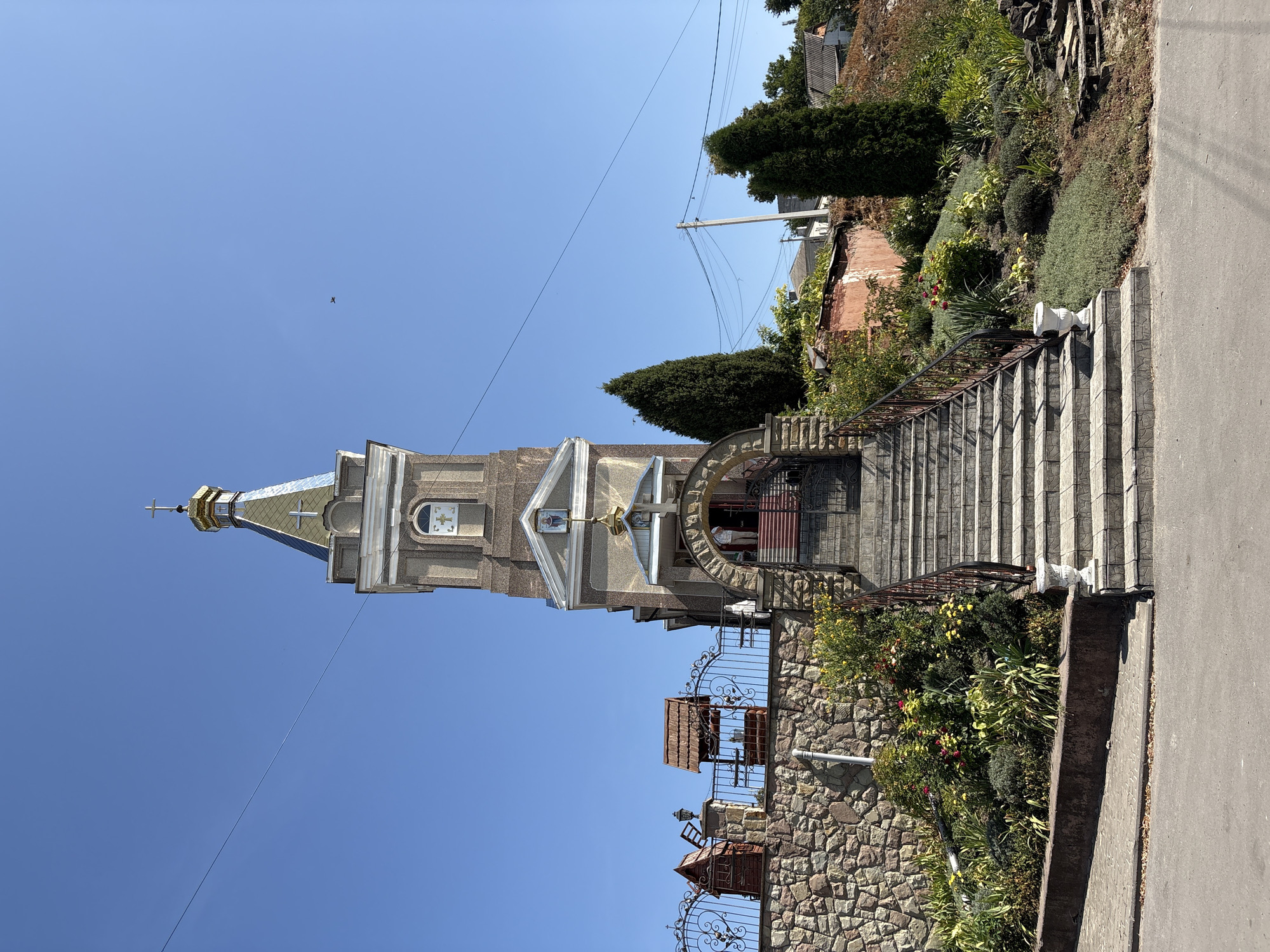 The image depicts the facade of a church, likely an Orthodox church given its architectural style, situated in the city of Lanivtsi, Ukraine. The church features a prominent bell tower with a golden dome and a cross at its apex. The tower also has a clock face. A set of wide, stone stairs leads up to the arched entrance of the church. The stairs are flanked by decorative railings and a well-maintained garden with a variety of plants and flowers. To the left of the stairs, a stone wall topped with an ornate metal fence encloses the church grounds. Visible on the grounds behind the fence are a small, decorative well and a brick structure resembling a miniature chapel or mausoleum. The sky is clear and blue, indicating a sunny day. The surrounding environment suggests a rural or suburban setting, with houses and trees visible in the background. There are no people clearly visible in the image, though a person can be partially seen through the entrance arch.