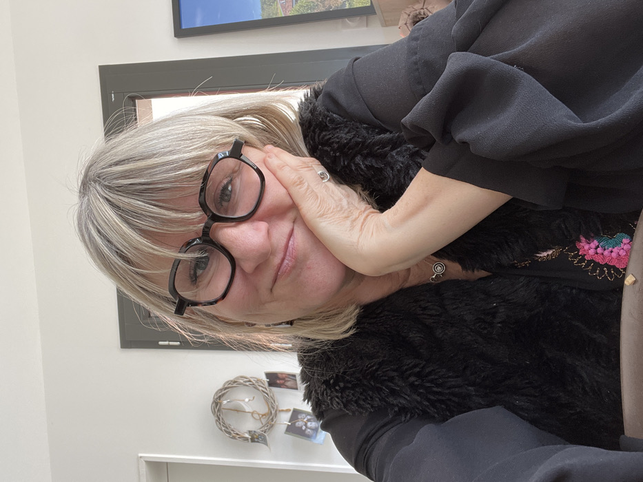 An adult female is depicted in a close-up, chest-up self-portrait taken indoors, located in Chambœuf, France. She has light-colored hair with visible gray roots/highlights, styled with bangs, and wears dark, angular-framed eyeglasses. Her expression is a slight smile with closed lips. Her left hand, adorned with a silver-colored ring on the ring finger, supports her head against her left cheek and chin. She is dressed in a dark long-sleeved garment, layered with a black faux fur vest, and wears a silver-colored circular pendant necklace. Below the necklace, an embroidered pattern featuring pink, green, and gold threads is visible on her attire. The background features a plain white wall. To the left, a woven wreath with small attached photographs is visible. Centrally behind her, a dark-framed door or window is present. To the far right, a framed image or screen displays a landscape scene. A small, light-colored object is positioned on a surface in the lower foreground.