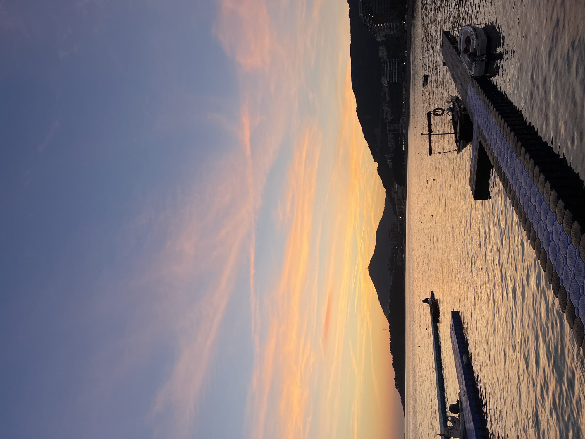 The image depicts a coastal scene at sunset in Rafailovići, Montenegro, with a bay foreground and silhouetted mountains in the background.
The sky occupies the upper two-thirds of the frame, displaying a gradient from deep blue at the top, transitioning through lighter blue to vibrant horizontal streaks of pink, orange, and yellow clouds near the horizon, indicative of a sunset.
The horizon is defined by two prominent dark landmasses. To the left, a smaller, low-lying headland is silhouetted. To the center-right, a larger, steep mountain dominates, also silhouetted in dark grey, featuring a small antenna-like structure at its peak. Beneath these landmasses, a strip of coastal development is visible, consisting of numerous dark building structures, some with visible illuminated windows or lights, extending along the shoreline.
The water in the bay reflects the warm sunset colors, appearing in shades of orange and yellow, with slight ripples on its surface. Several dark, small buoys are scattered across the water.
In the midground, two distinct floating modular docks are present. The left dock is constructed from blue and white plastic modules, extending from the left edge towards the center. A small, white motorboat with an outboard engine is moored to its left side. Further back along this dock, a small, dark boat with a single, indistinct, silhouetted figure seated or crouching aboard is moored. The figure faces right, observing the scene. The right dock, constructed from blue and dark grey/black plastic modules, extends from the bottom right corner towards the center-left. A dark, traditional-style boat featuring a mast-like structure and a white life ring is moored at its far end. Closer to the foreground on the right dock, a white dinghy or small motorboat is moored.