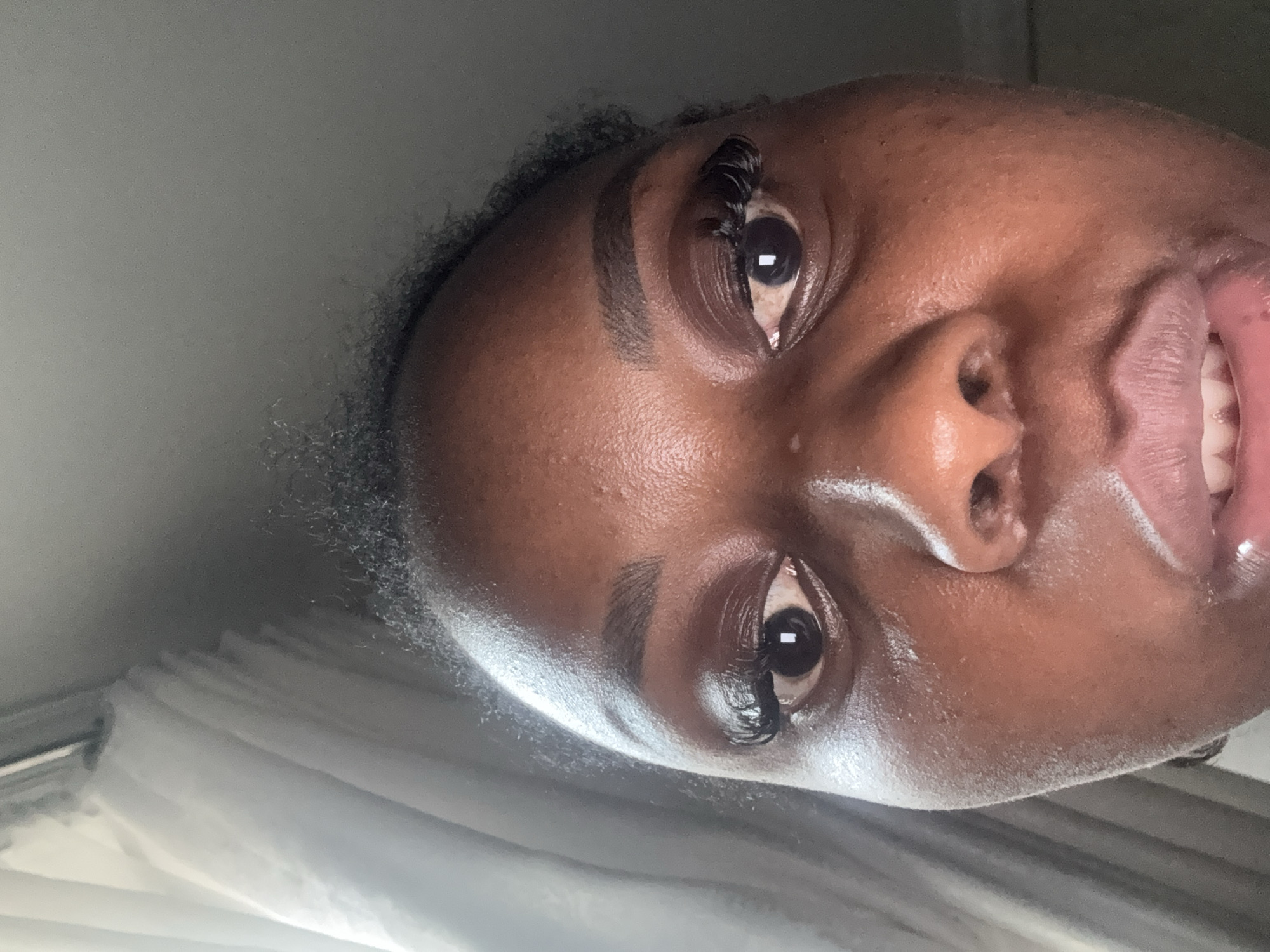 This media file is a close-up, rotated portrait of a person's face, focusing on their eyes, nose, and mouth. The person has dark skin and appears to be wearing makeup, with defined eyebrows and mascara on their eyelashes. Their eyes are open and looking towards the camera, and their mouth is slightly open, revealing teeth and a hint of a smile. The lighting suggests it might be daytime, possibly indoors, given the soft, diffused light and the blurry background which shows what appears to be white curtains or blinds. There is no visible text in the image. The overall impression is of a casual, self-taken photograph.