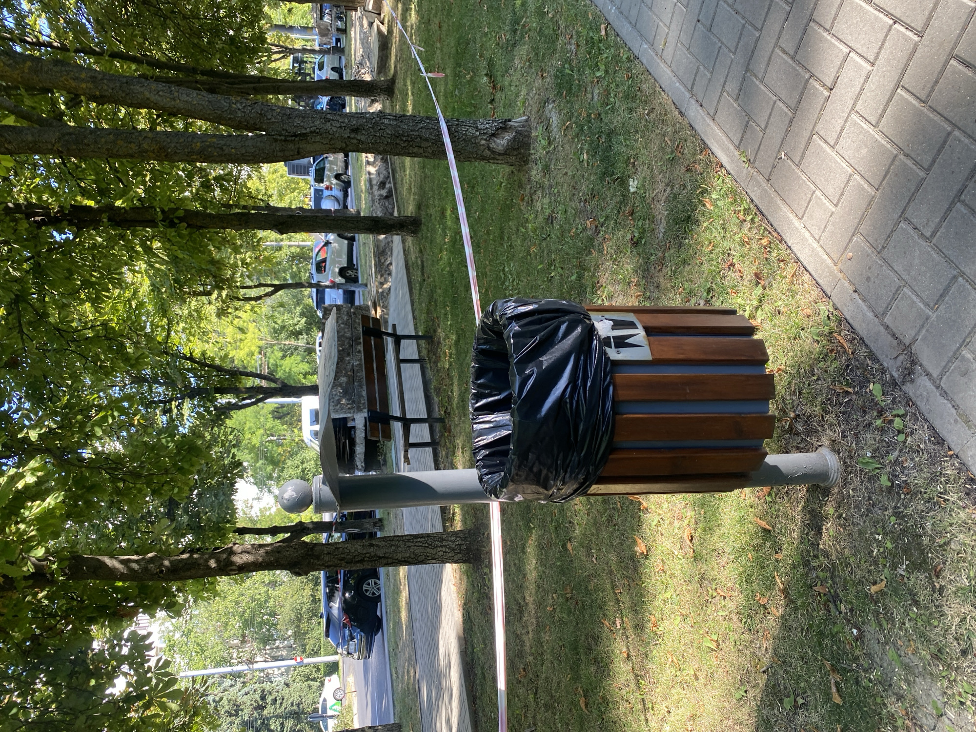 A public trash can with a black plastic bag liner stands on a grassy area next to a paved walkway. The trash can is made of wood slats and metal. Red and white caution tape stretches across the grass, indicating a restricted area. In the background, several cars are parked on a street lined with trees. A bench and a large, rough-hewn stone are also visible in the park-like setting. The scene suggests an outdoor urban environment, likely a park or sidewalk area within Chișinău, Moldova.
