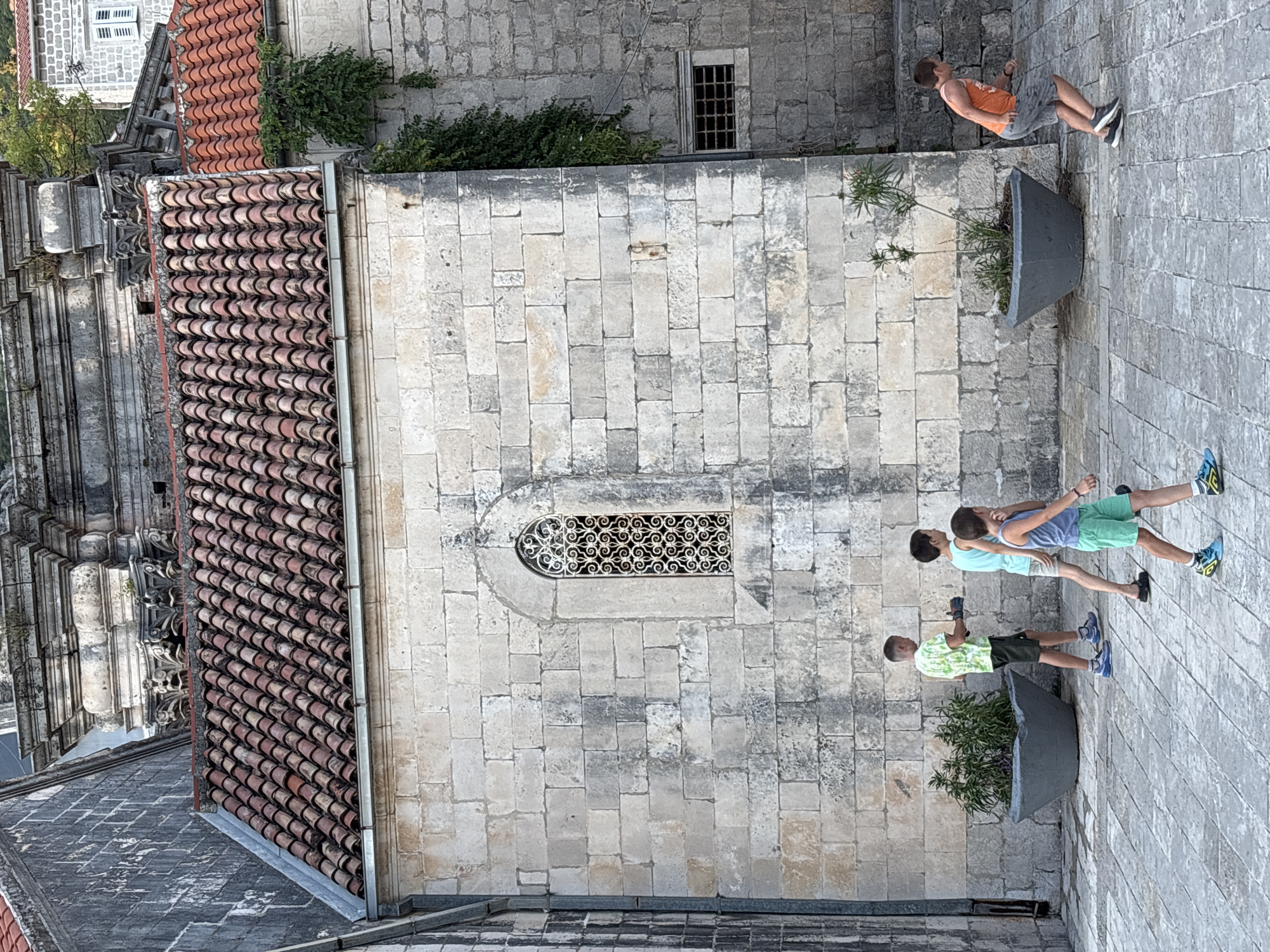 In the historic city of Perast, Montenegro, four young boys are walking on a paved courtyard. The boy in the foreground, on the right, wears a blue tank top and green shorts, with blue and yellow athletic shoes. To his left, another boy, wearing a light blue t-shirt and beige shorts with black shoes, walks with his head turned. Further left, a boy in a green and white patterned t-shirt and black shorts with blue shoes walks with his hands gloved. The fourth boy, partially visible on the far left, is also wearing athletic shoes. The boys are walking past large, grey, conical planters containing greenery. The background consists of an old stone building with a tiled roof and a decorative arched window with intricate metalwork. Additional stone structures and greenery are visible further back, suggesting an old European setting. The lighting suggests it is daytime.