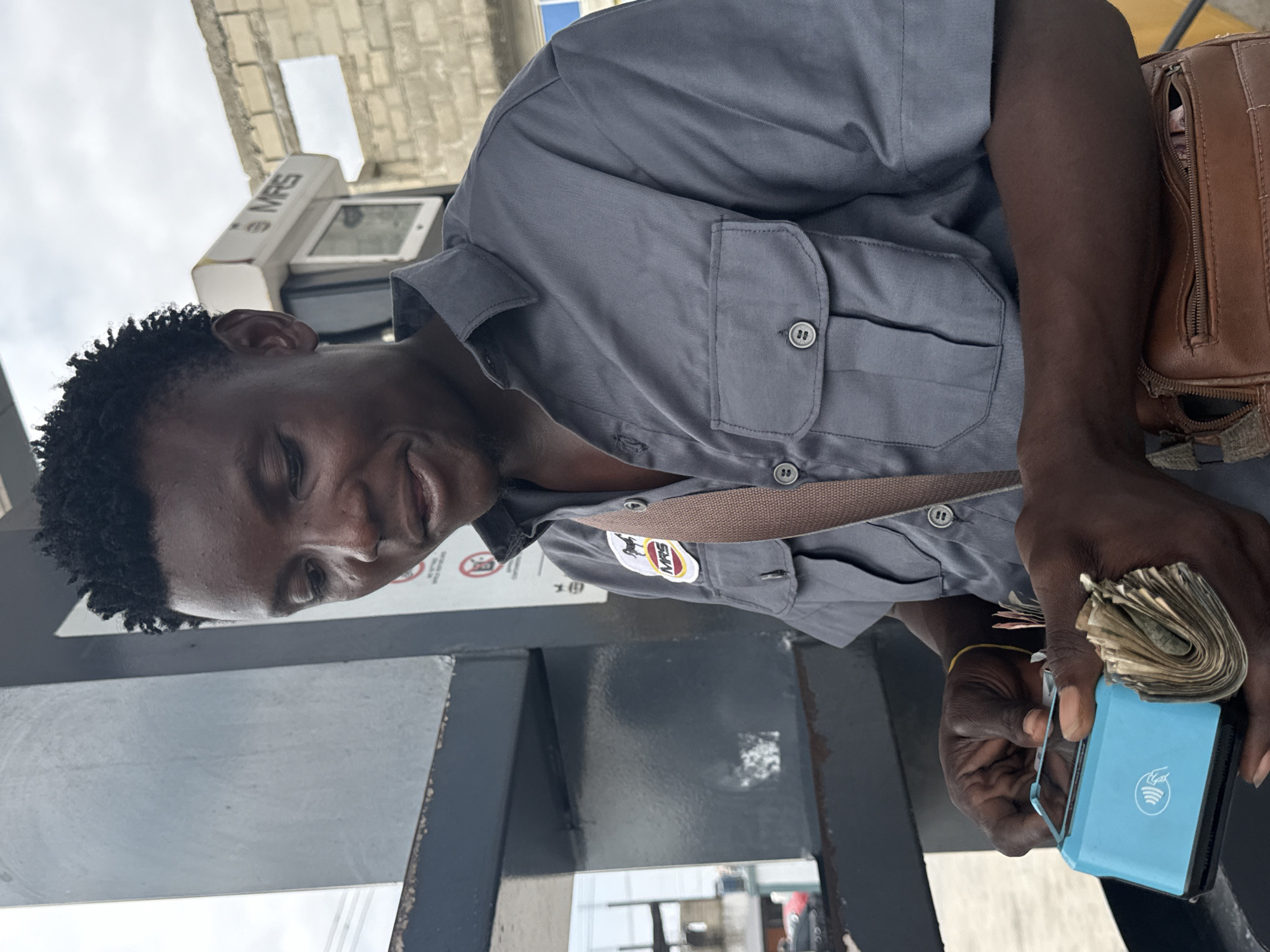 In Ado, Nigeria, a dark-skinned man wearing a grey button-up shirt with a circular MRS logo on the left breast is standing outdoors, likely at a gas station given the visible fuel pump and brick wall in the background. He is holding a light blue card reader in his left hand and a wad of cash in his right hand. The card reader has a contactless payment symbol. The man appears to be in the process of conducting a transaction, possibly collecting payment from a customer. A brown leather satchel is slung across his body. The lighting suggests it is daytime.