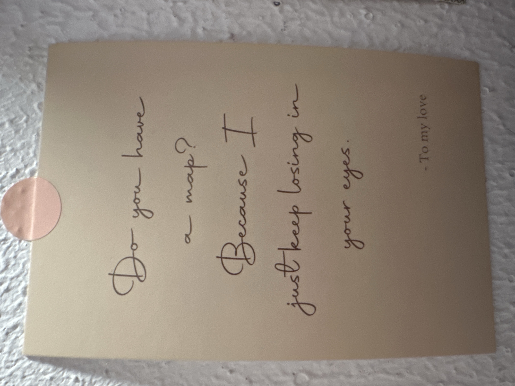A light brown or beige card with dark brown text is prominently displayed, tilted to the left. A round, light pink sticker is affixed to the top left corner of the card. The card rests on a textured, off-white surface, possibly a wall or fabric, with soft shadows creating some depth. The lighting suggests an indoor setting.
The text on the card, written in a decorative, flowing cursive script, reads:
"Do you have
a map?
Because I
just keep losing in
your eyes.
- To my love"
The message is a romantic or affectionate pick-up line or sentiment, signed "To my love," indicating a personal and emotional connection. There are no people visible, and no specific activity is depicted beyond the presentation of the card itself. The overall impression is one of a thoughtful, intimate gesture.