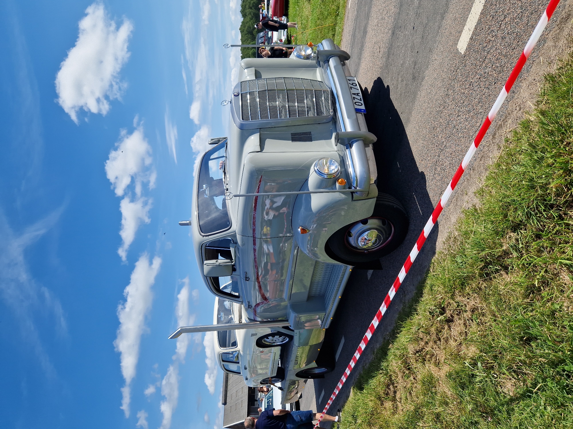 In Edenberga, Sweden, on a sunny day with scattered clouds, a vintage grey Mercedes-Benz truck is parked on an asphalt road, its trailer carrying a vintage white car. The truck, a classic model with a prominent chrome grille and twin headlights, has a tall exhaust stack on its side. A red and white striped ribbon runs along the grassy roadside, indicating a boundary. In the background, several people are visible, some standing and observing, while others are near parked cars, suggesting a car show or event. The environment is rural, with fields of green grass and trees in the distance. The license plate of the truck reads "OZA 761".