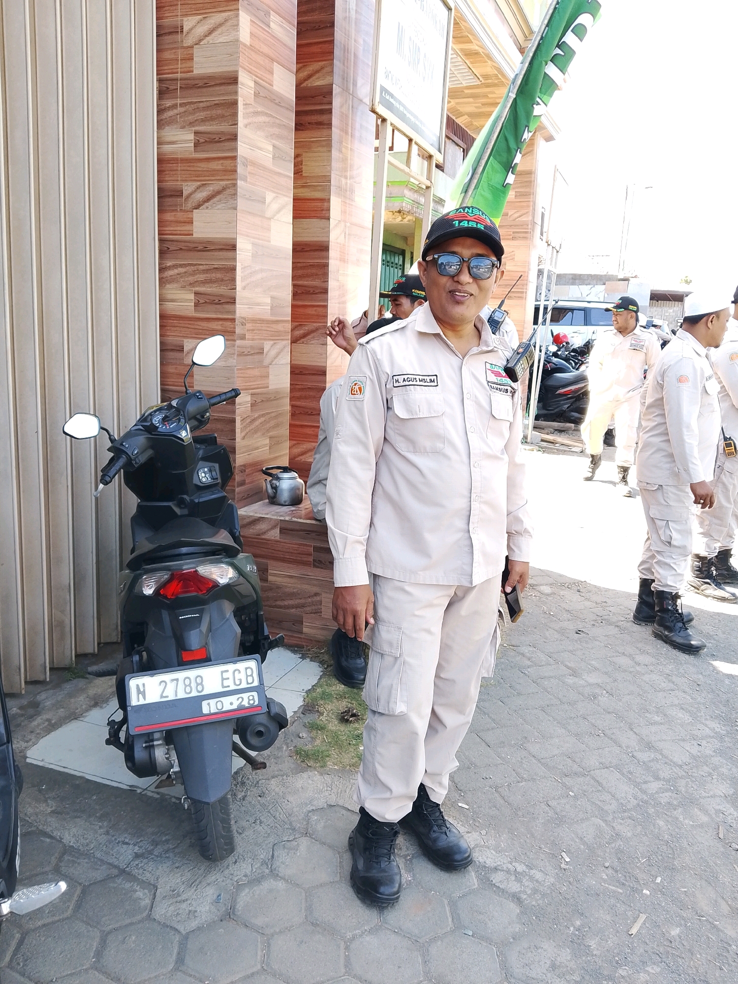 In Banjarejo, Indonesia, a man wearing a tan uniform, sunglasses, and a black cap stands on a paved surface. He is positioned next to a black scooter with a license plate that reads "N 2788 EGB 10.28". The man has a small radio clipped to his uniform. Behind him, a building features a tiled facade with a light-colored vertical stripe pattern. Several other individuals in similar tan uniforms are visible in the background, some holding flags. The scene is outdoors, under bright sunlight.