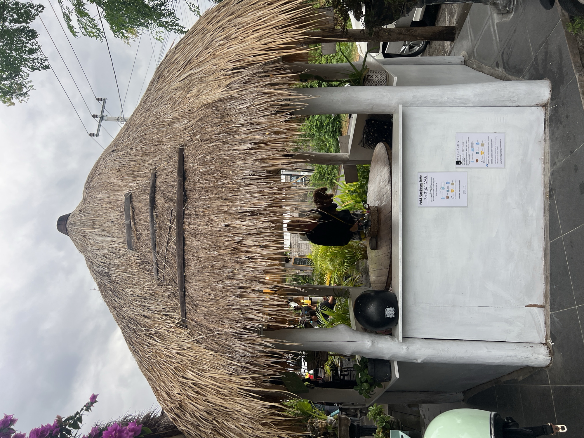The image captures an outdoor pavilion with a thatched roof, serving as a "Peduli Alam Sorting Station" in Abang, Indonesia. A person, partially visible, stands behind a wooden counter, interacting with something on the counter. A black helmet rests on the counter, to the left of the person. The pavilion is supported by white-painted pillars and features a white counter base. Two informational posters are affixed to the counter base, one titled "Peduli Alam Sorting Station" and dated "30 JULY 2026," and another titled "What do I do with my trash?" and "PEDULI ALAM RECYCLING CENTER." Lush green plants and foliage surround the structure, suggesting a tropical or garden setting. In the foreground, a light green scooter helmet is visible on the left. The sky is overcast with clouds. The overall scene depicts a functional sorting station, likely for waste management, integrated into an outdoor public space.
